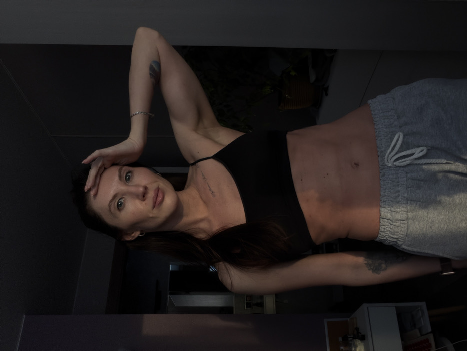 A female individual is depicted indoors in what appears to be a residential setting located in Perm, Russia. The subject, an adult with long dark hair, light-colored eyes, and fair skin, is positioned facing the camera. She is wearing a black spaghetti strap crop top or sports bra and light grey drawstring sweatpants, revealing her midriff with visible abdominal musculature. Her right hand is raised to her forehead, fingers resting on her temple, while her left arm hangs at her side. A silver-colored bracelet is visible on her right wrist, and a dark-colored smartwatch or fitness tracker is on her left wrist. Small hoop earrings are present in both ears. A subtle tattoo is visible on her upper left chest, and a darker mark or tattoo is on her left bicep.
The scene is lit from the left, casting warm light on the subject's left side and creating shadows on her right side and much of the background. To the left of the subject, a light-colored multi-drawer unit or nightstand is visible, displaying small items including a white container and a white rectangular object. Further left, a doorway or opening leads to a darker interior. To the right of the subject, a window sill or shelf is discernible, holding a plant in a terracotta-colored pot and other indistinct objects in shadow. The wall behind the subject to the left appears to be a muted purple or dark grey.