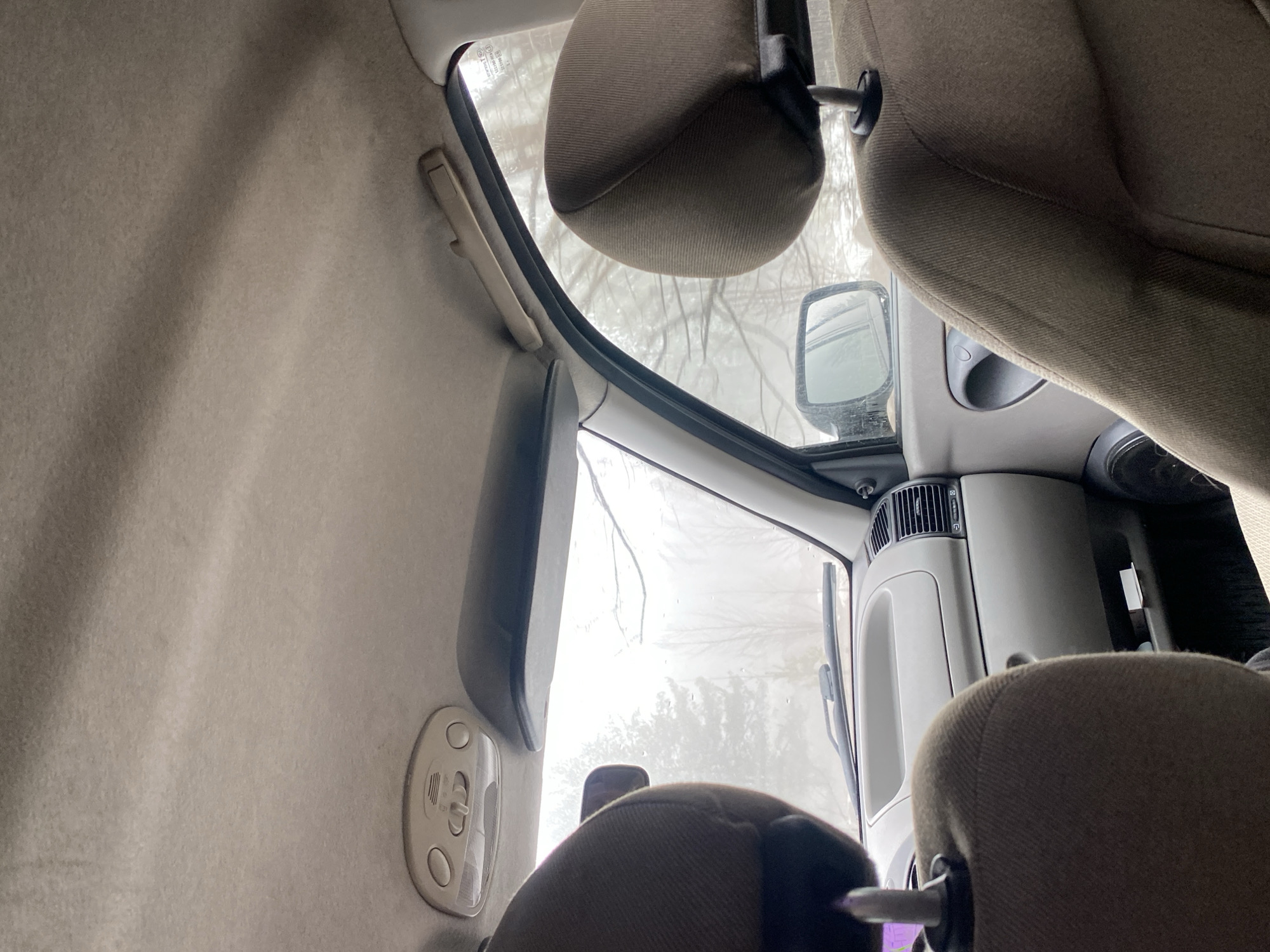 The interior of a vehicle is visible, likely a van or a larger passenger vehicle. The focus is on the front area, with the windshield showing a blurred view of a snowy or overcast outdoor environment. Bare tree branches are visible through the windshield, suggesting a winter or early spring season. The vehicle's interior includes upholstered seats with headrests, a rearview mirror, a sun visor, and interior lighting. The dashboard or console area is partially visible, featuring air vents and storage compartments. The overall interior color scheme is neutral, with light beige or gray tones.
Given the prompt, the location is presumably Chaikovsky, Russia, though this cannot be definitively confirmed from the image alone.