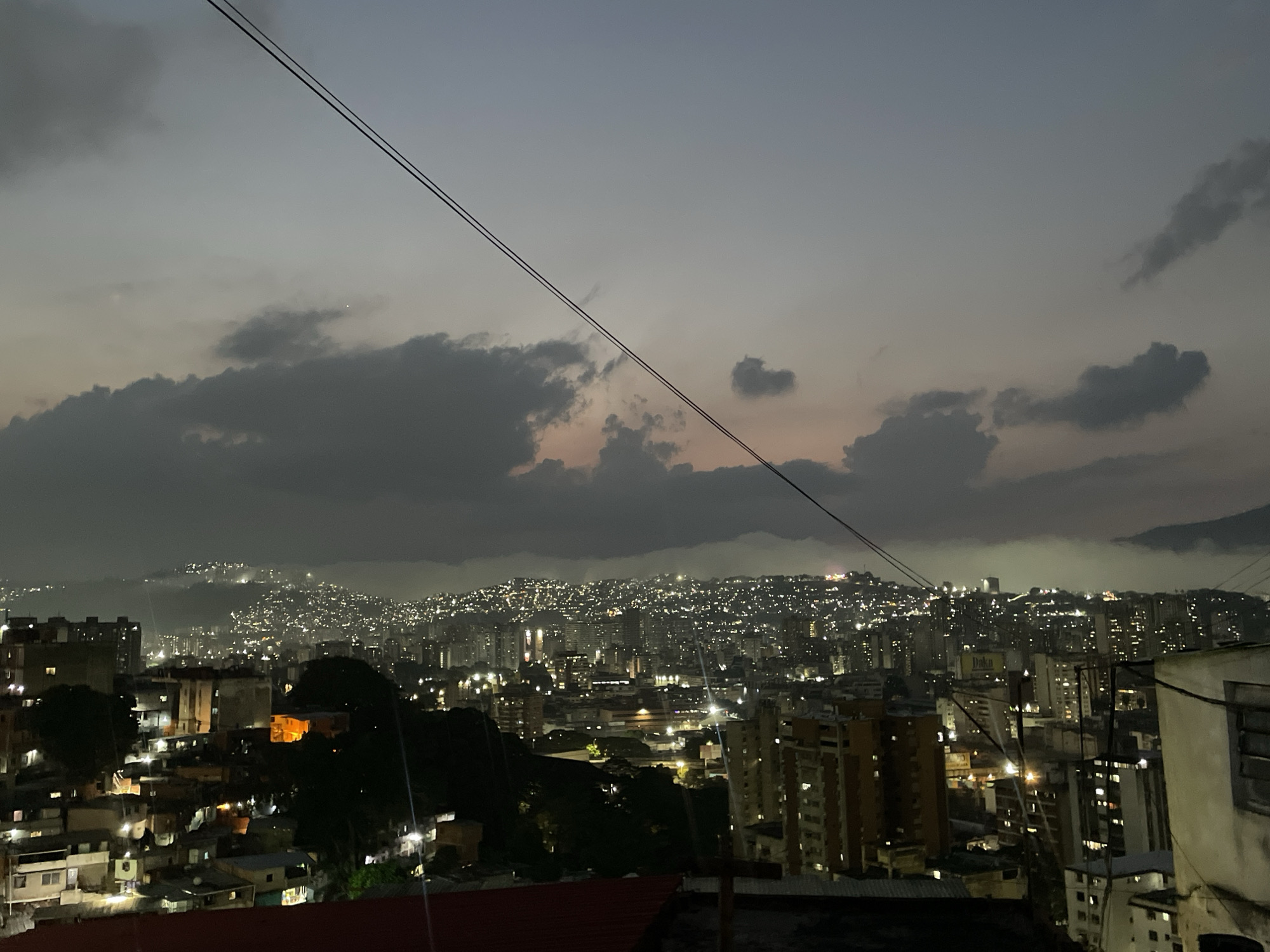 The image shows a nighttime view overlooking the city of Caracas, Venezuela. The urban landscape is densely populated with buildings of varying heights, illuminated by streetlights and lights from within the structures. In the distance, the hills surrounding the city are also dotted with lights, indicating settlements on the slopes. Overhead, a cloudy sky is visible, with two power lines stretching across the frame. The foreground features a residential area with rooftops and buildings of a smaller scale compared to the high-rise structures in the central view. The overall scene conveys an urban environment at dusk or early night.