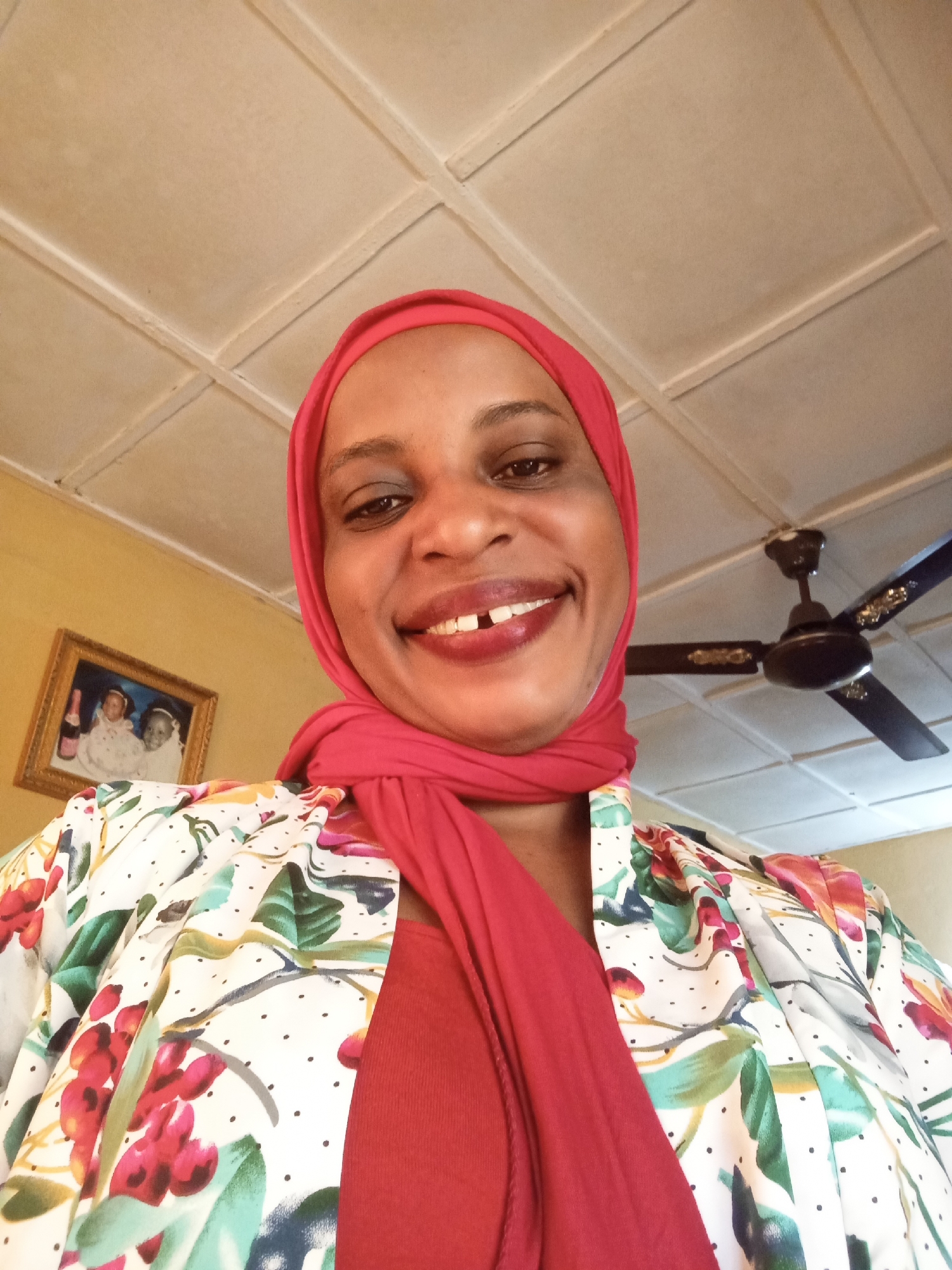 A close-up, low-angle selfie shows a Black woman wearing a red hijab and a floral print jacket over a red top. She is smiling, revealing slightly uneven teeth. In the background, a framed picture hangs on a yellow wall, and a black ceiling fan with decorative elements is visible. The ceiling is white and paneled. The image appears to be taken indoors. This scene is located in Narayi, Nigeria.