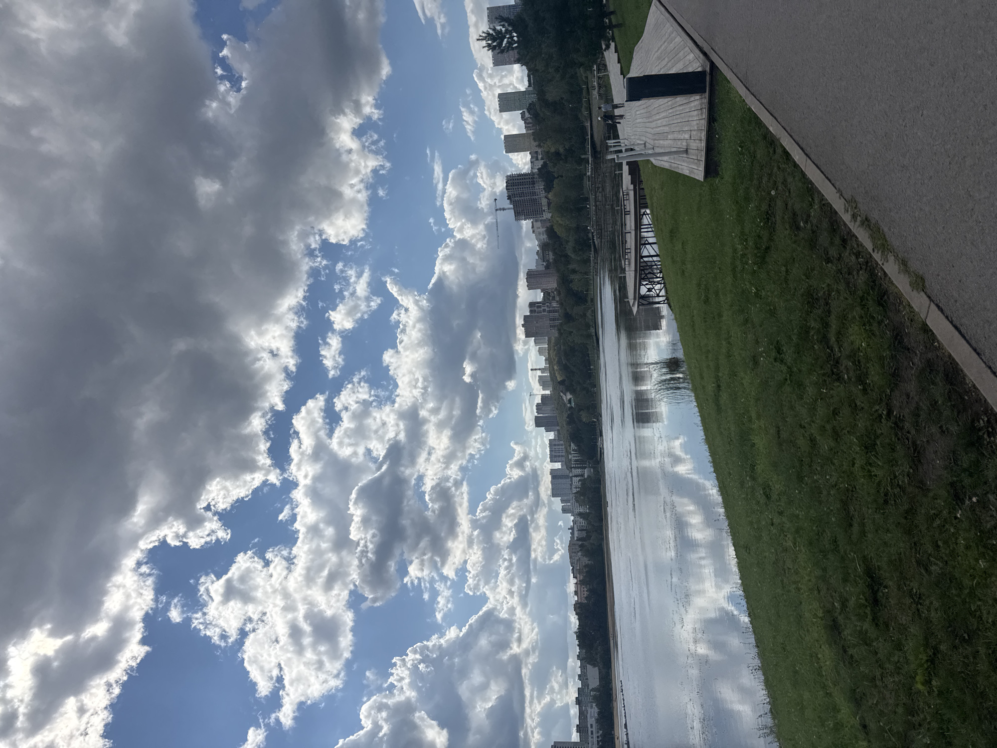 This image shows a scenic view of a lake and a city skyline in Ufa, Russia. The foreground features a paved walking path with a grassy embankment leading down to the water. The lake is calm, with reflections of the sky and clouds visible on its surface.
In the middle ground, there is a wooden pier with several tiered platforms, and a few people can be seen walking on it. The background is dominated by the city of Ufa, with a mix of residential buildings and some under-construction structures, including a crane. Lush green trees line the edge of the lake between the water and the city. The sky is partly cloudy with dramatic, fluffy white clouds interspersed with patches of blue sky, suggesting a dynamic weather day. The overall atmosphere is peaceful and urban, showcasing the blend of nature and city life.