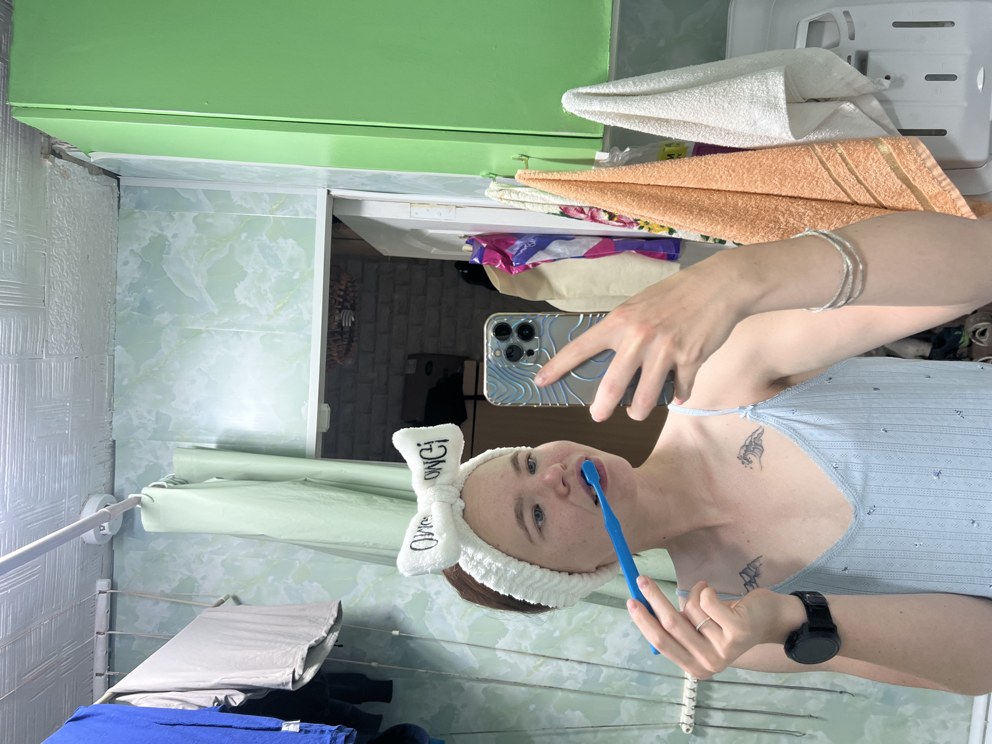 A young Caucasian woman with light skin and auburn hair, wearing a white fuzzy headband with a bow that reads "OWGI OWGI" and a light blue camisole, brushes her teeth with a blue toothbrush. She is holding a smartphone in her right hand, reflecting the image in a mirror. She has two tattoos on her chest: one of a mountain range on the left and a wave on the right. She is also wearing a black smartwatch on her left wrist and a silver bracelet on her right arm. The background appears to be a bathroom with light green patterned wallpaper, a clothesline with several items of clothing hanging, a green cabinet, and several towels hanging on hooks. The room is part of a residential building, possibly a bathroom in an apartment in the city of Chaikovsky, Russia, given the context provided. The lighting is dim and appears to be artificial.