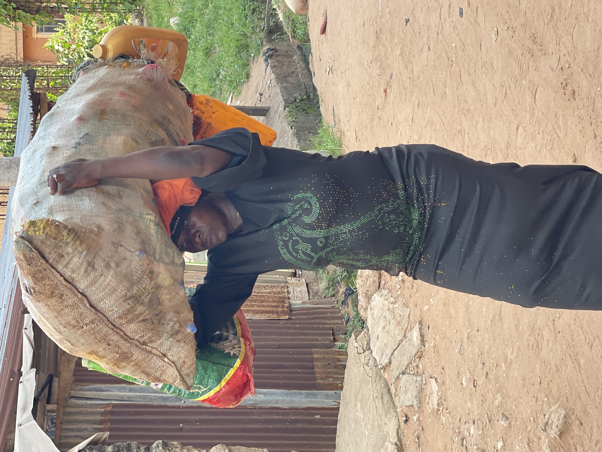 In Nkpor, Nigeria, an adult woman is seen carrying a large burlap sack filled with an assortment of recyclables, including a yellow plastic container. The woman wears a dark-colored long dress with green sparkling patterns. She supports the heavy sack with one arm across the top and another arm around the side. The location appears to be near a building with corrugated metal walls and in a dirt/sandy environment. Patches of green vegetation are visible in the background.