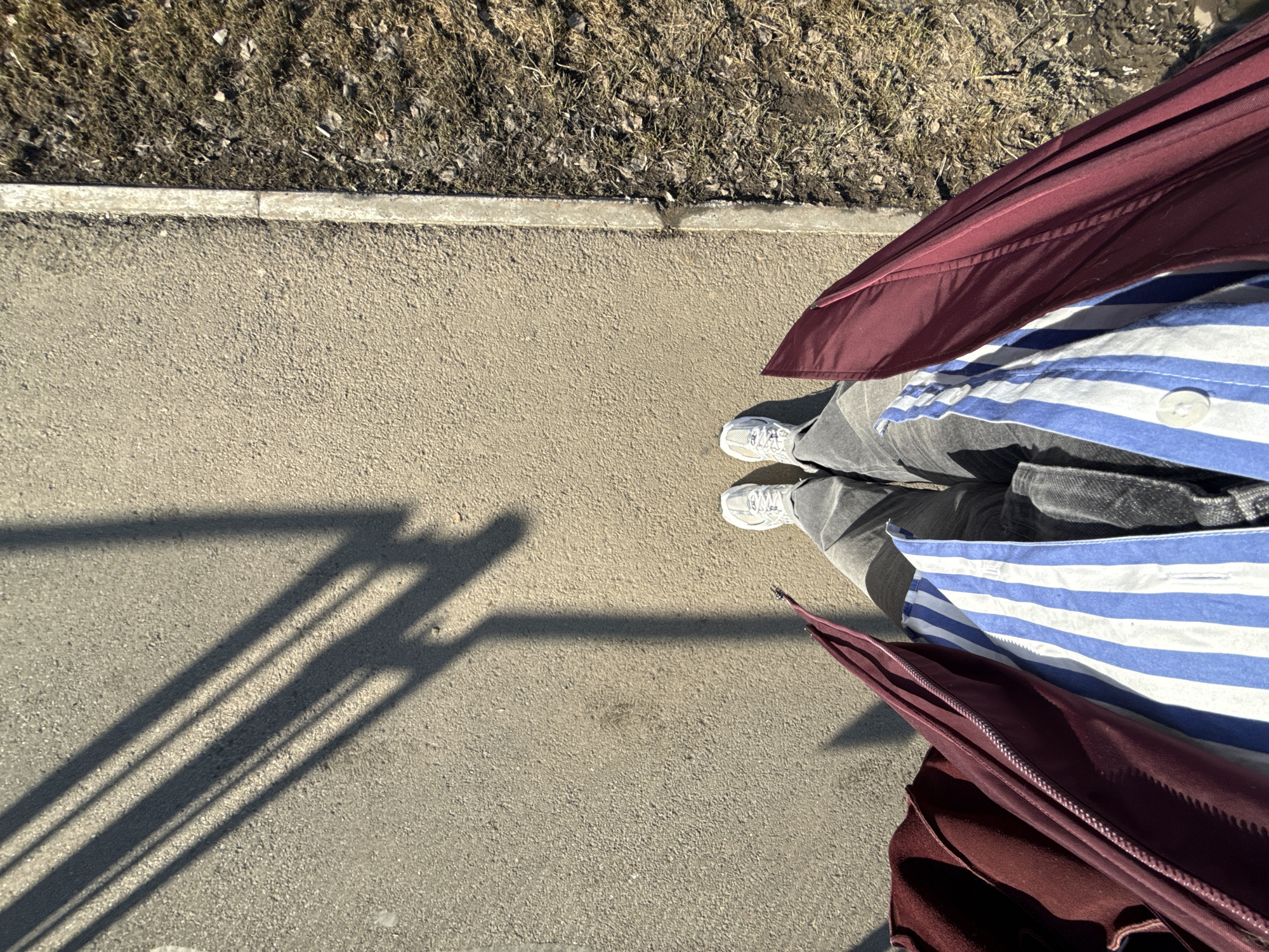 The image presents a view from above, looking down at a section of a pavement in Izhevsk, Russia. The pavement is concrete, with a curb bordering a patch of dry grass and soil. To the right, a person's legs and feet are visible. They are wearing grey sneakers, grey jeans, a blue and white striped shirt, and a burgundy jacket. A shadow of a structure, possibly a fence or railing, is cast on the pavement to the left. The scene is brightly lit, suggesting a sunny day.