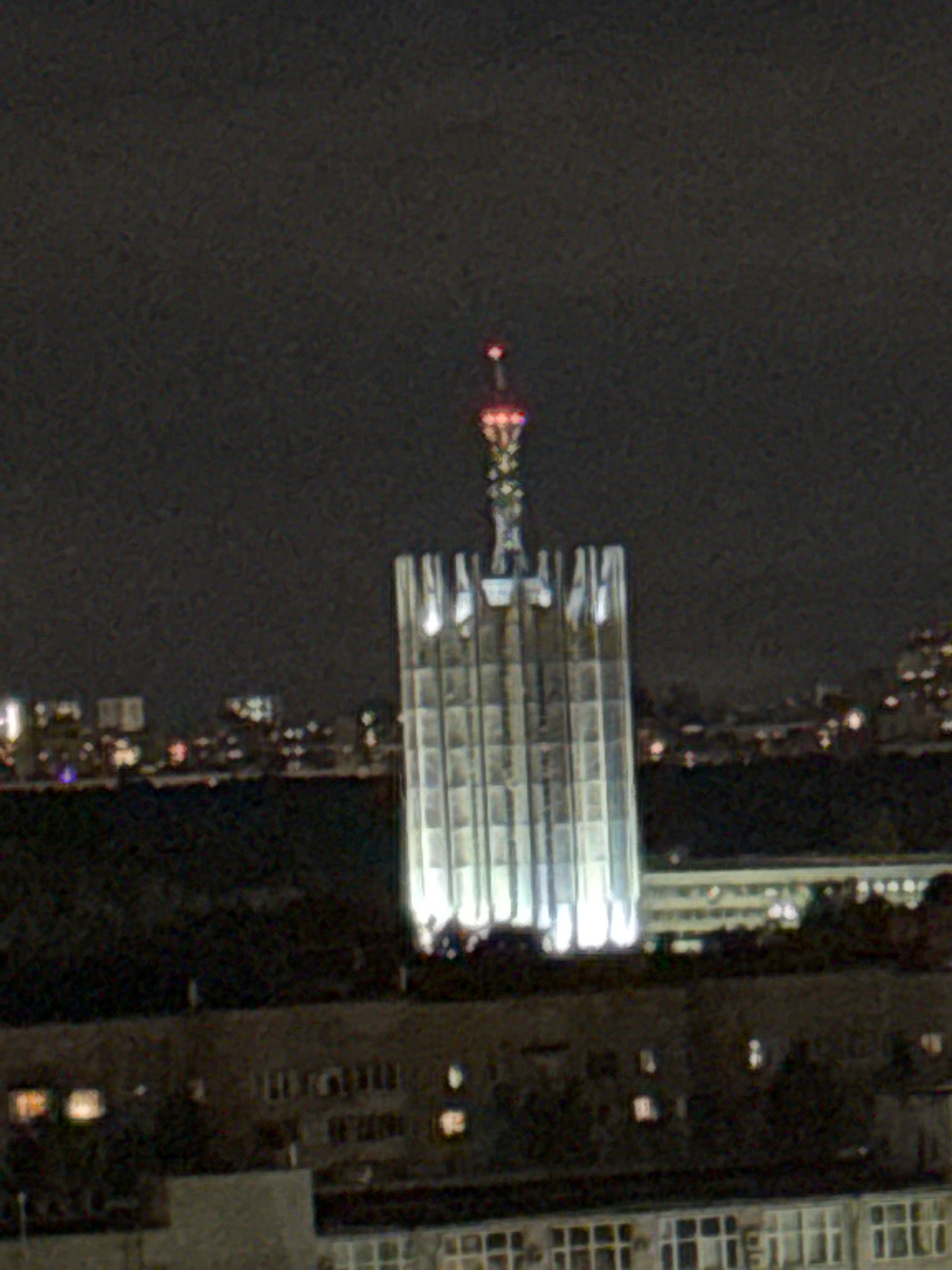 The image shows a nighttime view of a tall, illuminated building with a spire at its peak. The building is a dominant structure in the scene, with a distinct architectural style characterized by vertical lines and illuminated panels. In the foreground, lower buildings with visible windows are present, suggesting a cityscape. The overall environment is dark, indicating it is nighttime, and the sky is a uniform dark gray. The lighting on the main building is bright and white, contrasting with the darker surroundings. There are no people visible in the image. The location is identified as Plintovka, Russia.