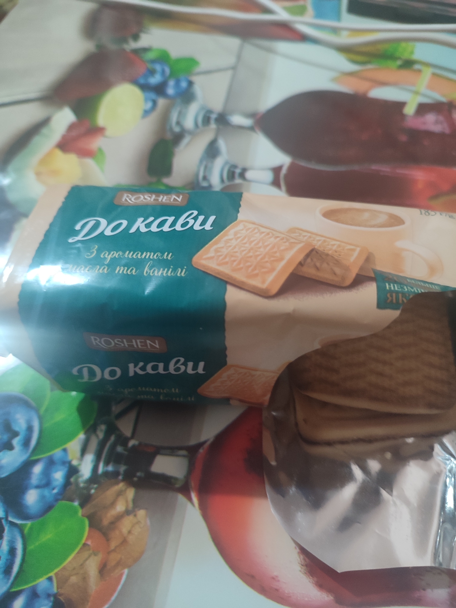 A package of Roshen "Do кави" cookies with coffee flavor, cream flavor, and vanilla flavor is on a table. The cookies are rectangular and light brown in color with a patterned surface. The packaging is primarily teal and white, with images of blueberries and fruits. The scene appears to be indoors, possibly in a kitchen or dining area, in Chernivtsi, Ukraine. The lighting is artificial and somewhat dim, casting shadows on the table and the cookies. The image is taken from a slightly elevated angle, looking down at the package.