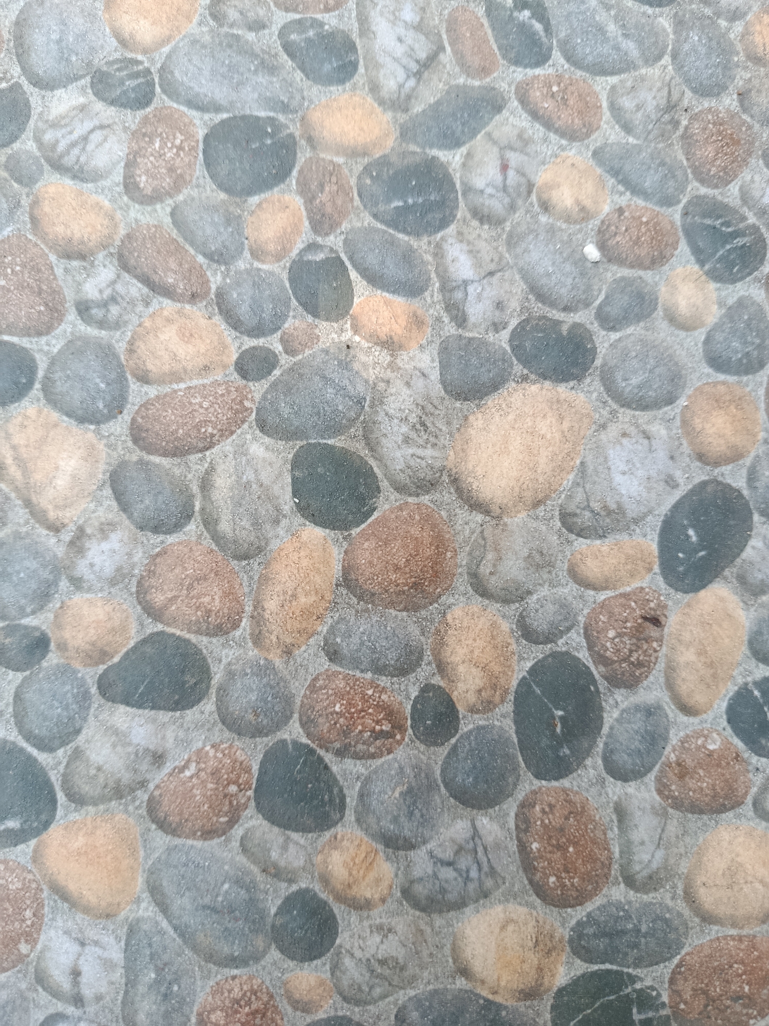 The image shows a close-up, top-down view of a pebble tile floor. The pebbles are rounded and vary in size, color, and texture, with shades of gray, brown, and tan. The tiles are arranged in a mosaic pattern with visible grout lines between the pebbles. The overall impression is one of natural earth tones and a textured surface. No people or specific locations in Wanasari, Indonesia are visible in this abstract image of a flooring material.