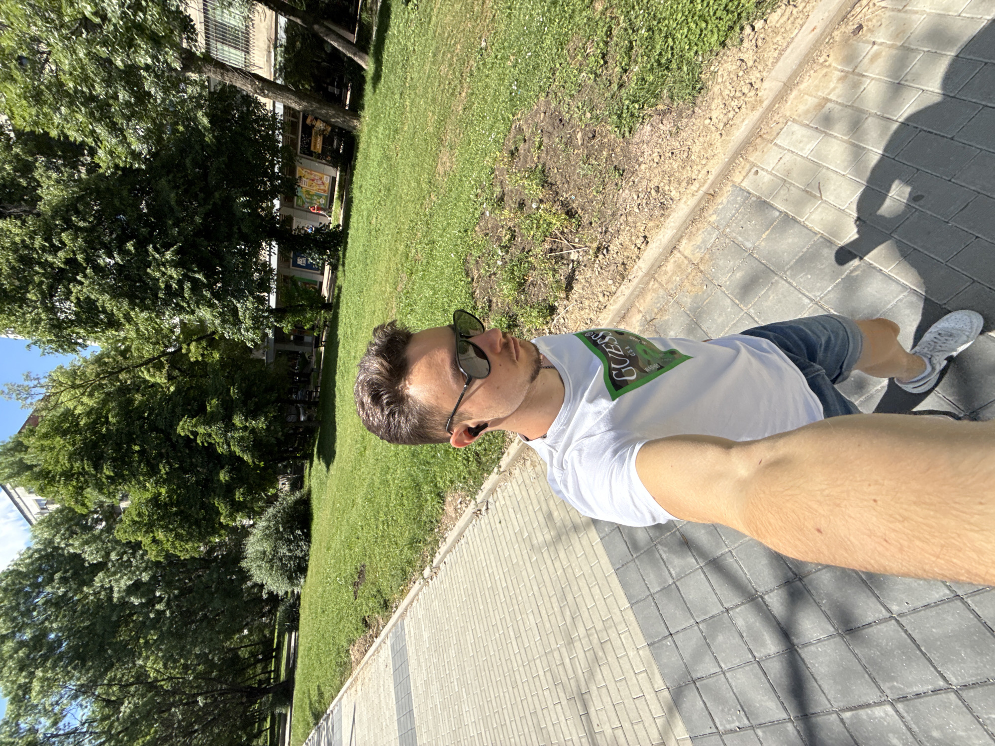 A person stands on a brick-paved walkway in Sofia, Bulgaria. They are taking a selfie, holding their arm outstretched. The person wears a white t-shirt with a "Guess" logo, denim shorts, and grey sneakers, and they have sunglasses on. To the left is a grassy area with trees, and in the background, buildings are visible. The scene is well-lit, indicating a sunny day.