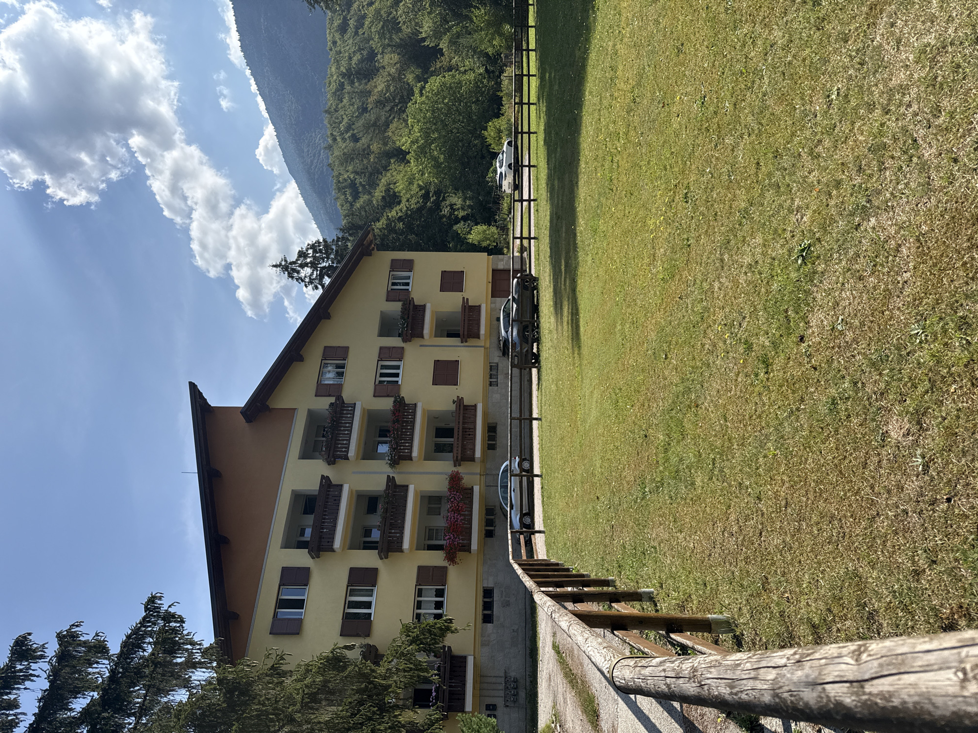 The image shows a yellow apartment building with multiple balconies in San Lorenzo Dorsino, Italy. The building is situated in a grassy area bordered by a wooden fence. Three cars are parked in front of the building, and a dense green forest covers the hillside behind it. The sky is blue with scattered clouds.