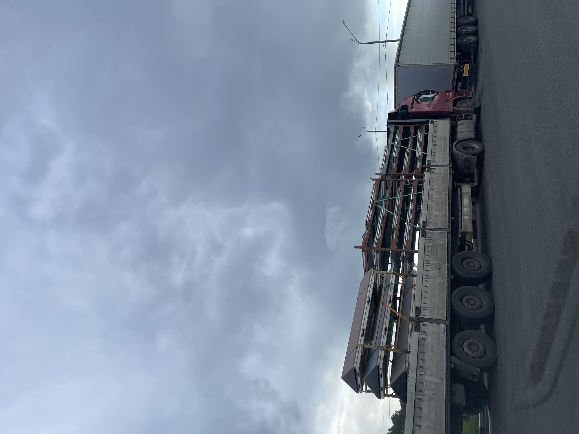 The image shows a semi-trailer truck carrying a load of what appear to be prefabricated building components in Бавыкино, Russia. The truck is positioned on an asphalt road under an overcast sky. The load is secured with straps and consists of multiple large, flat panels supported by a metal frame. Another truck, a semi-trailer type, is positioned behind the first one. Streetlights are visible in the background.