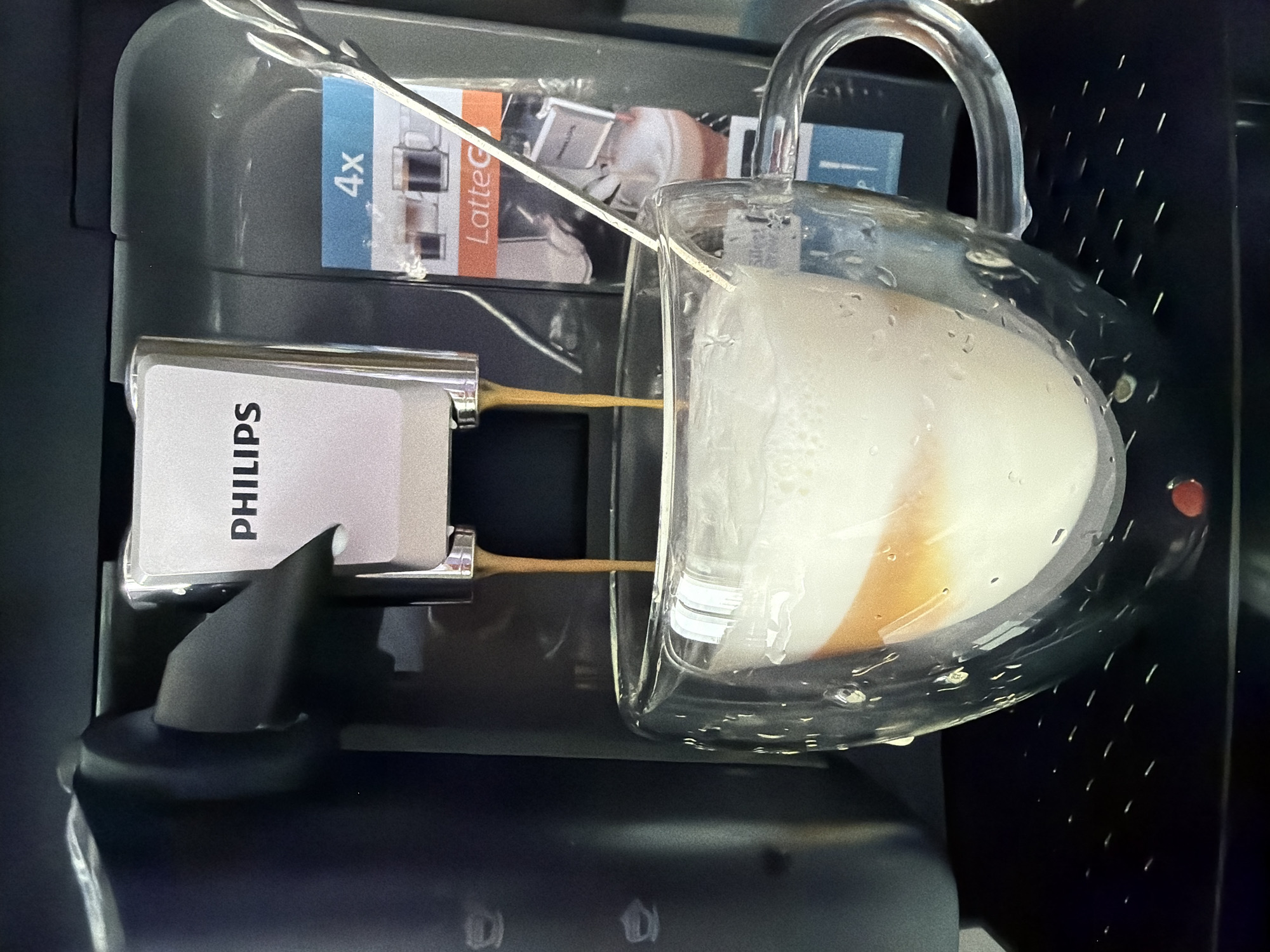 A Philips LatteGo coffee machine is brewing a latte into a clear glass mug. The machine is black and gray with a Philips logo on the side. The mug is filled with coffee and steamed milk, showing distinct layers. A silver spoon is also visible. The scene is indoors, but there is no information to verify the location to Chaikovsky, Russia.