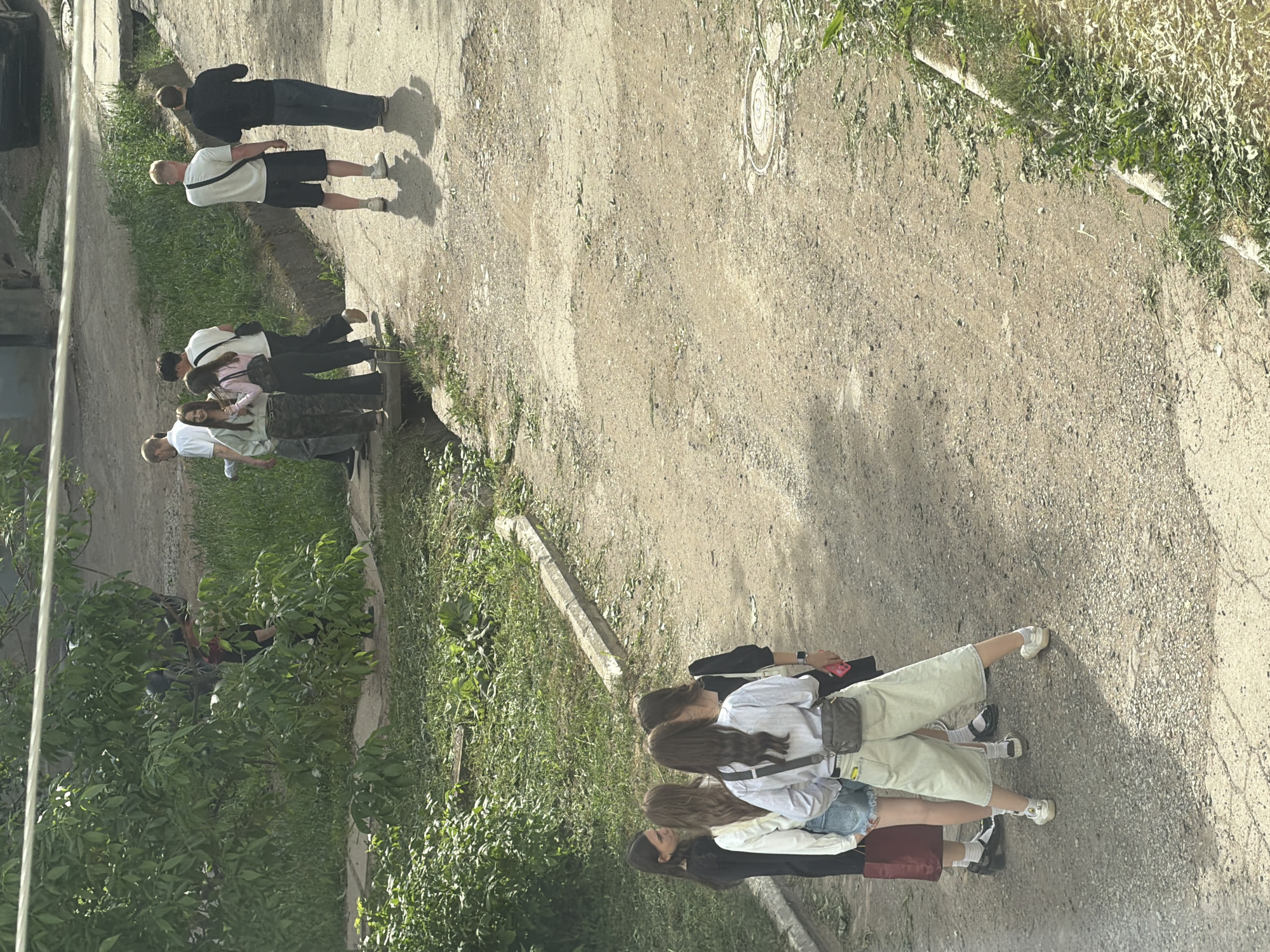 A medium-angle shot shows a group of people walking along a dirt road adjacent to a green space; the setting is Votkinsk, Russia. In the foreground, a group of four young women, dressed in casual summer clothes, walk together; their hair is long and brown. Further down the dirt road, a group of two men and one woman walks; a man, woman and child walks slightly away from the other group. In the distance, two men walk together. The dirt road runs parallel to a green space containing trees and grass. A manhole cover is visible. The scene takes place on a bright, sunny day, indicated by the shadows cast by the individuals.
