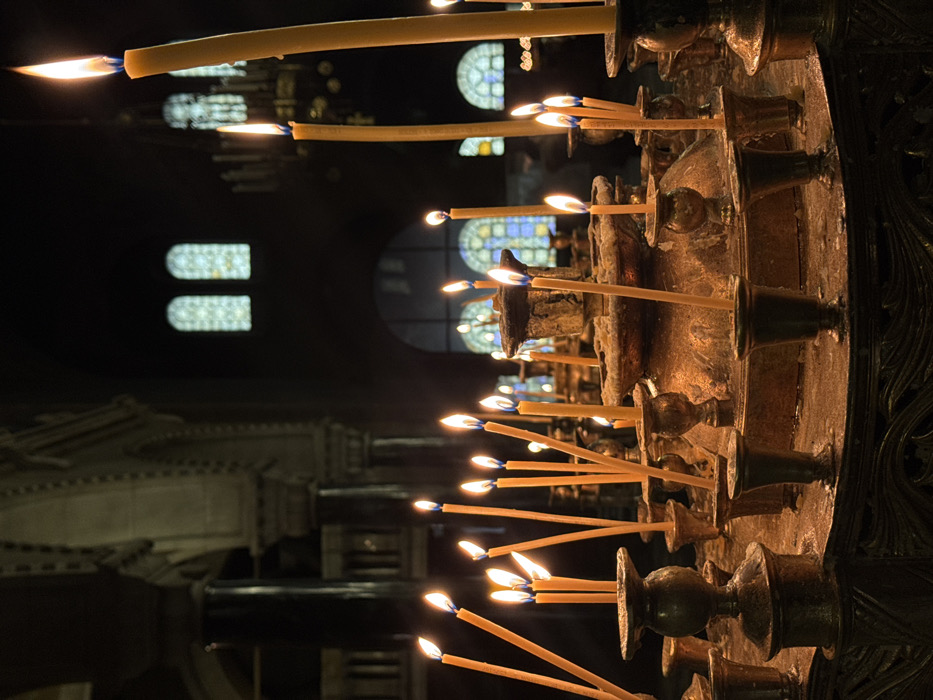 The scene depicts a dimly lit interior within the St. Alexander Nevsky Cathedral in Sofia, Bulgaria. In the foreground, a prominent, multi-tiered candelabra, appearing to be crafted from aged brass or bronze, is heavily encrusted with dried wax. It supports numerous thin, yellow beeswax candles of varying heights; most are actively burning, exhibiting bright, elongated flames that cast a warm, orange-yellow light. Additional, similar candelabra with lit candles recede into the midground. The background reveals the cathedral's grand architecture, including large, dark, ornate columns, a series of archways, and distant, arched stained-glass windows displaying geometric patterns, through which faint, diffuse light penetrates. A large, ornate chandelier is partially visible in the upper right background. No individuals are present, but the multitude of lit candles indicates recent human activity, likely for prayer or ceremonial purposes.