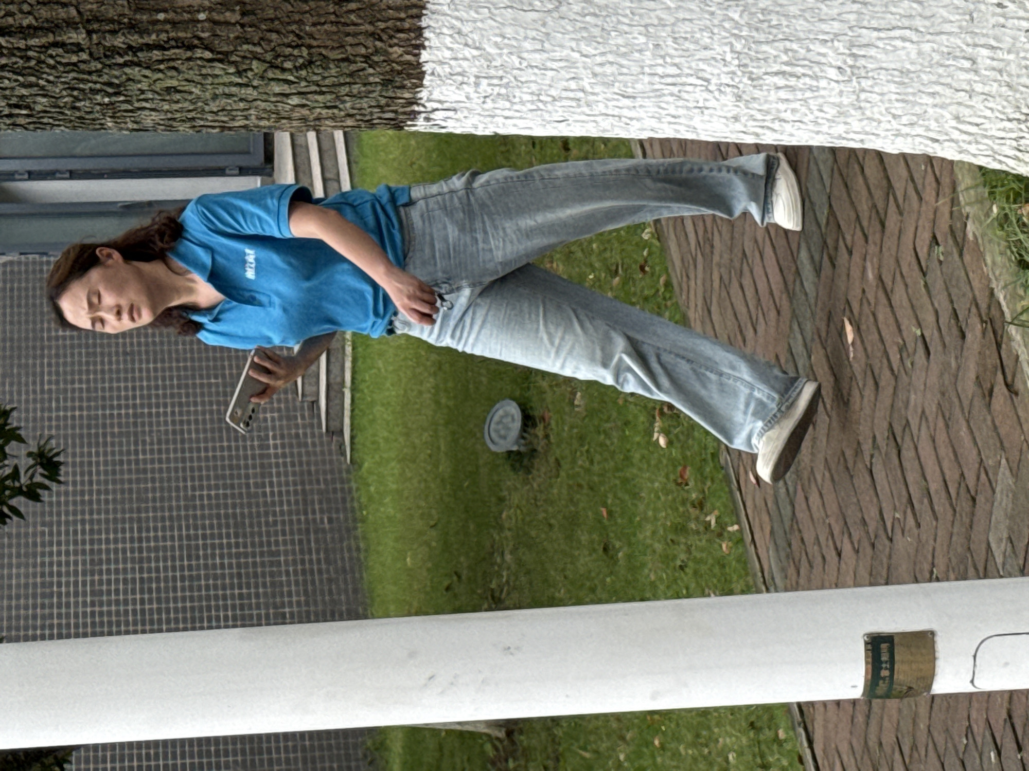 A woman with long brown hair is standing on a paved walkway holding a cell phone. She is wearing a blue t-shirt with white lettering and light blue jeans. She appears to be looking down at the phone. The location appears to be in Jiangjin, China, based on the style of brickwork on the walkway, the type of tree trunk, and the style of the signage on the pole at the bottom of the image. The environment includes a tree with white paint on the lower portion of its trunk, a grassy area, and a gray tiled wall or walkway.