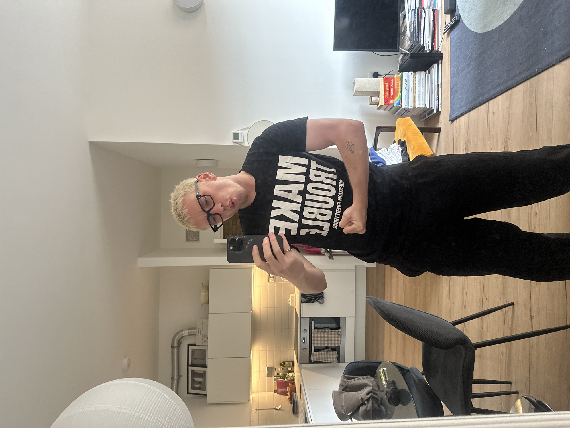 A man with bleached blonde hair and black-rimmed glasses poses in front of a mirror, taking a selfie with a black smartphone. He's wearing a black t-shirt with white lettering that reads "MAKE TROUBLE ABOUT." He is wearing black pants. The man is flexing his right arm, showing a tattoo. The background suggests an interior space, possibly an apartment, with a kitchen visible behind him. There are white cabinets, a tile backsplash, and a stainless-steel oven. A dining table with a gray chair is in the foreground. Other items, like books, a TV, and a rug, are in the background. The location appears to be an apartment interior in London, United Kingdom, based on the architectural style and interior design elements.
