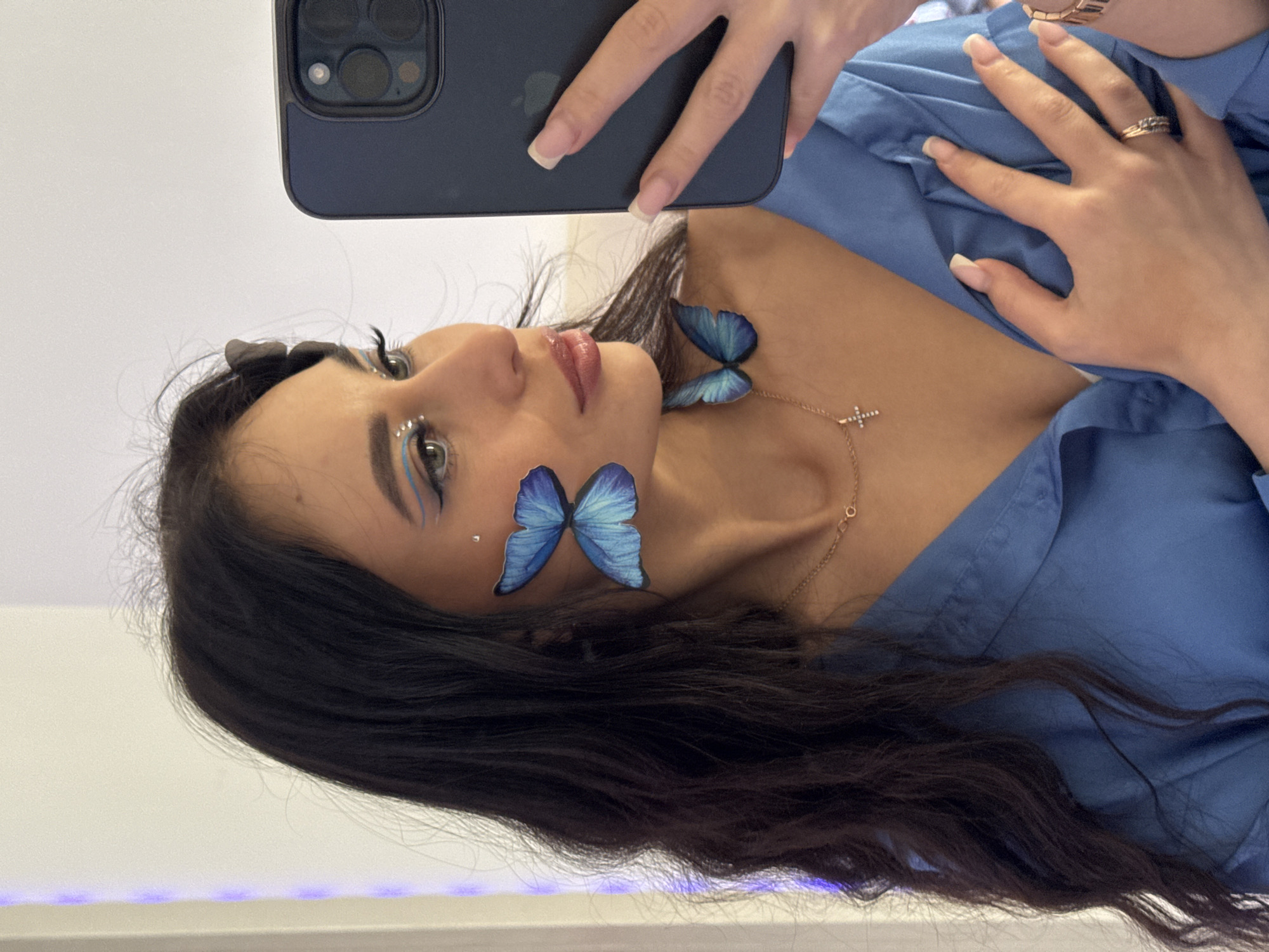 A woman with long, dark, wavy hair is holding a dark-colored smartphone in her right hand to take a selfie. She is wearing a light blue button-down shirt. She has makeup on, including blue eyeliner and small, shiny decorations around her eyes. There are blue butterfly decorations on her face and collarbone. She is wearing a delicate gold necklace with a small cross pendant. She is also wearing rings on her fingers and a watch on her wrist. The background appears to be a plain white wall. There is no mention of the location in the city of Фоки, Russia.