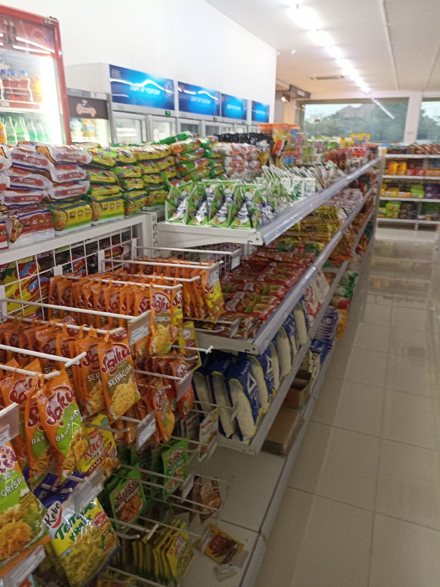 The image shows the interior of a convenience store. The store is well-stocked with various food items, primarily packaged snacks and instant noodles, displayed on shelves and in hanging racks. The shelves are organized in aisles, and the floor is tiled. In the background, refrigerators are visible, some of which are displaying beverages. The lighting in the store appears to be fluorescent. The overall impression is that of a typical retail environment in Indonesia, specifically in the city of Tompokersari, given the context. There are no people visible in the image, and the focus is entirely on the merchandise.