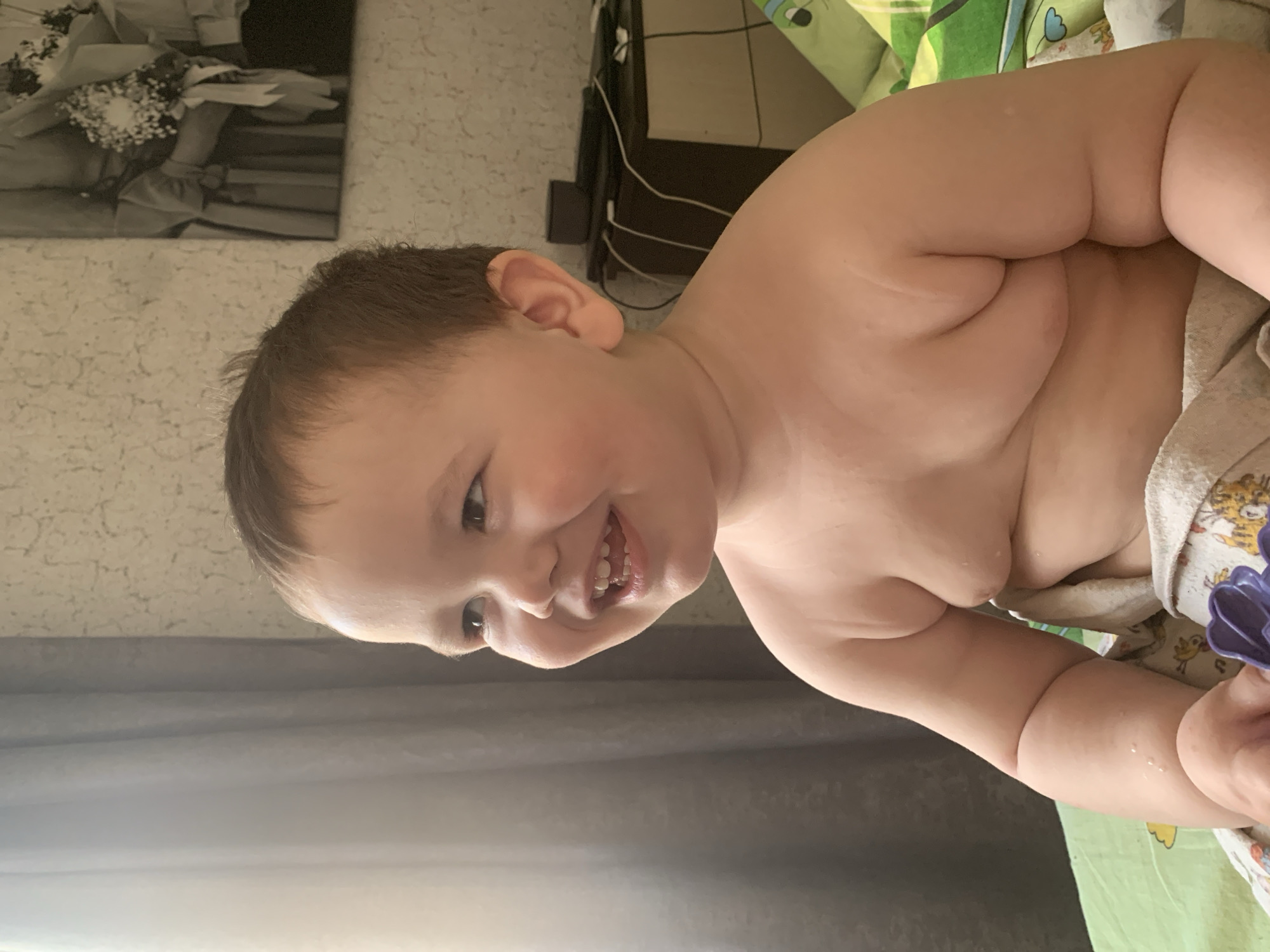 A close-up, tilted shot shows a fair-skinned baby with brown hair and a toothy grin. The baby is partially unclothed, revealing chubby arms and torso. A patterned blanket or towel is draped around the baby. The background includes a wall with patterned wallpaper, a black and white framed picture, and a bedside table with visible wires. The setting appears to be indoors, likely a home. There's no direct information to suggest this image was taken in the city of Фоки, Russia.
