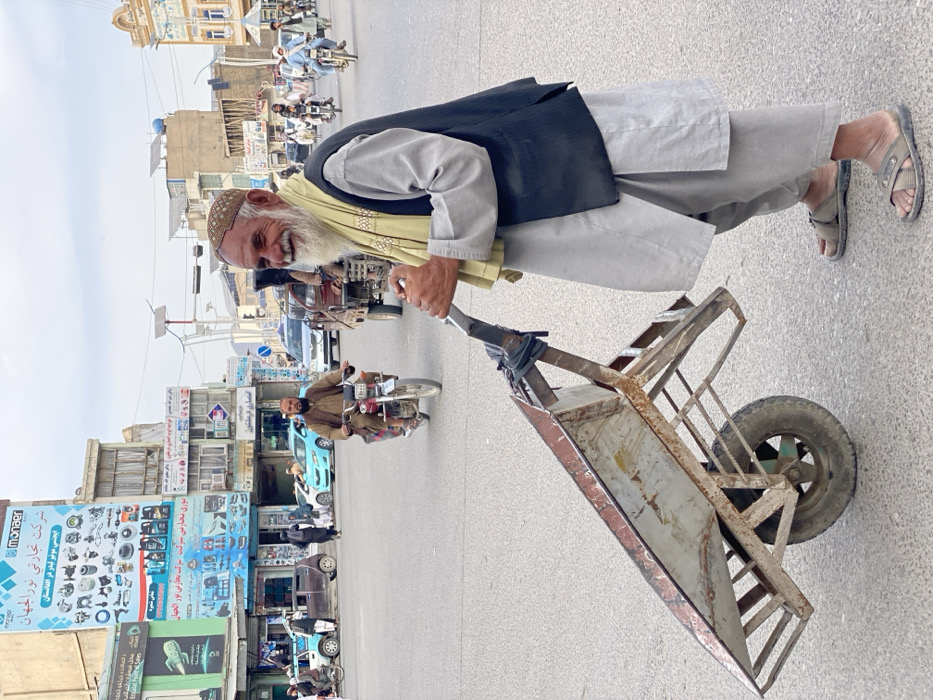 A smiling, elderly man with a long white beard and a patterned brown kufi pushes a single-wheeled, rusty metal wheelbarrow on a paved street in Kandahar, Afghanistan. He wears a light gray shalwar kameez, a dark ribbed vest, and a light yellow patterned scarf draped around his neck. His feet are clad in dark open-toed sandals. The wheelbarrow features a rectangular, deep basin and a metal frame.
The scene is a busy urban intersection. In the background, multiple men are riding motorcycles, while cars and other vehicles traverse the street. Multi-story buildings line the intersection, some appearing commercial with storefronts and various signage. A prominent blue and white billboard on a building to the left displays "Monaer" and "شرکت تجارتی نورالجهان" (Noorul Jahan Trading Company) with images of automotive parts such as oil filters and lubricants. Other individuals are visible as pedestrians and commuters. The sky is overcast and pale gray, and solar panels are visible on the rooftops of several buildings in the distance.