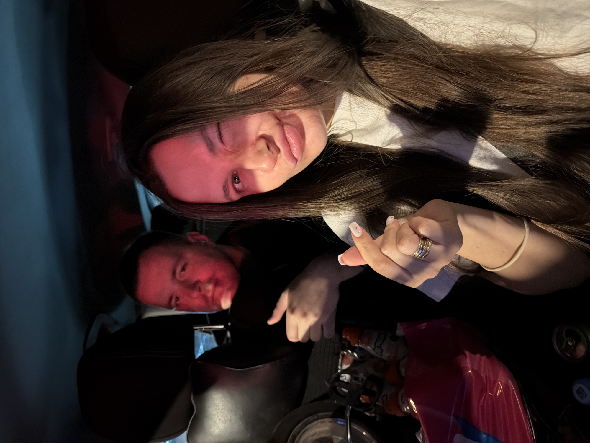 Two individuals, a man and a woman, are seated in the back of a vehicle, possibly a car or van. The man, on the left, is wearing a dark shirt and is partially obscured by the headrest of the front seat. He is looking forward, and his left arm is raised, with his thumb visible. The woman, on the right, is in the foreground, facing the camera. She has long, dark brown hair and is wearing a white t-shirt. Her face is illuminated by a red light, which casts a strong glow on the left side of her face. She is winking with her left eye and puckering her lips. Her right hand is raised, forming a heart shape with her thumb and index finger, and she is wearing rings on her fingers and a bracelet on her wrist. Behind the woman, a portion of the vehicle's interior is visible, including the back of the front seats and what appears to be some bags and bottles in the space between the seats. The scene is dark, with the red light being the primary source of illumination for the woman's face. There is no indication of the specific location within the city of Moscow, Russia.