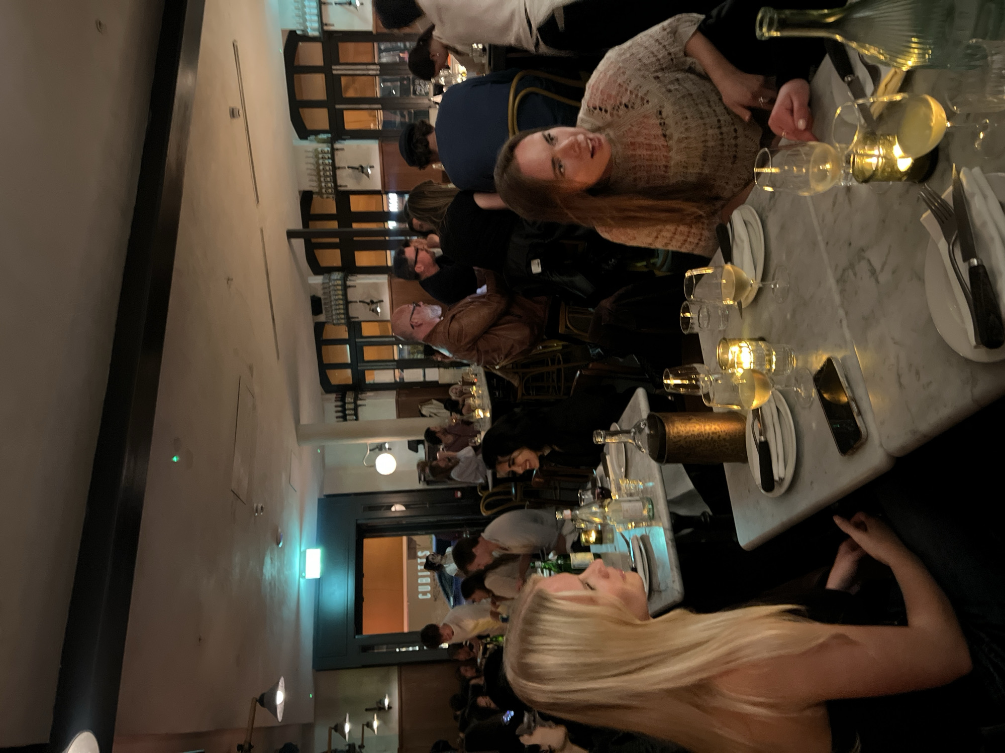 The image depicts a busy restaurant scene, likely in London, United Kingdom. Numerous patrons are seated at tables, dining and socializing. A woman with long blonde hair is in the foreground, facing the camera. Behind her, other diners can be seen, including a woman with long brown hair. The tables are set with plates, cutlery, glasses, and wine bottles. Some tables have decorative candle holders. A sign for "CUBITT'S" can be seen in the background. The restaurant has a modern decor with dark wooden fixtures.