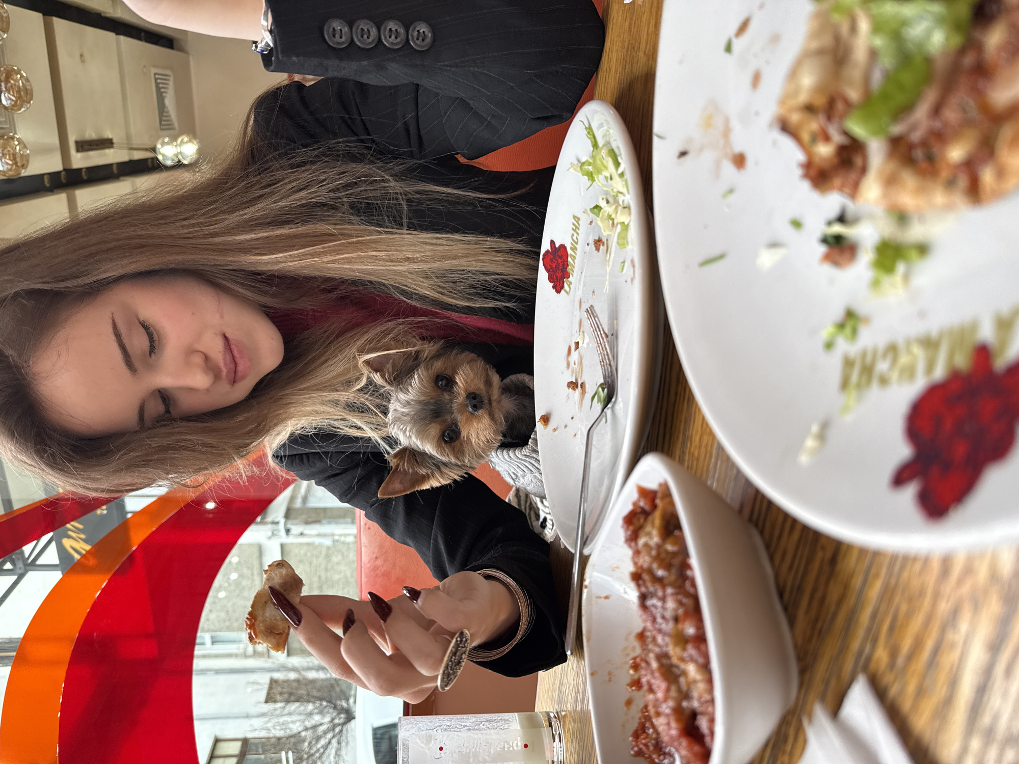 A young woman with long blonde hair is sitting at a wooden table at a restaurant, possibly in Sofia, Bulgaria. She is wearing a black pinstripe blazer over a red top. Nestled beside her is a Yorkshire Terrier dog, looking directly at the camera. The woman is holding a piece of food in her hand. There are several white plates on the table, some with remnants of food, suggesting a meal is underway or recently concluded. A small white bowl containing a tomato-based dish is also visible. The plate closest to the woman has the name "LA MANCHA" and a red flower printed on it. In the background, a window with red and orange panes is seen, along with an exterior view of buildings, hinting at an urban location. A decorative light fixture is slightly visible in the top left corner. The photo is taken from a close-up, slightly angled perspective.