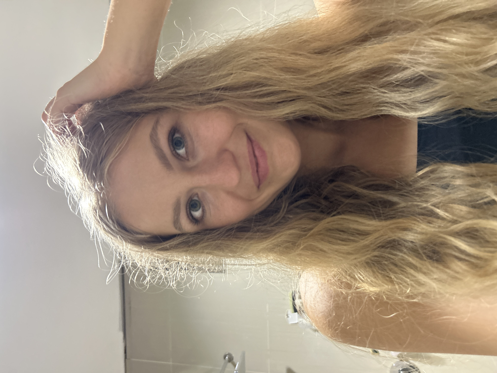 A young woman with long, wavy, golden-blonde hair is captured in a selfie-style portrait. Her head is slightly tilted, and she is looking directly at the camera with a gentle, friendly smile. Her blue-green eyes are prominent. Her left hand is visible, resting casually on the top of her head, touching her hair. She has fair skin and appears to be wearing a dark-colored top.
The background indicates an indoor setting, likely a bathroom, with plain white or light grey tiled walls. A chrome fixture, possibly a faucet or part of a sink, is partially visible in the lower section of the frame. The lighting is bright and even, illuminating her features and hair.
The woman is posing for the self-portrait with a relaxed and pleasant demeanor. There is no visible text within the image. The provided context for the location is Kuta Selatan, Indonesia.