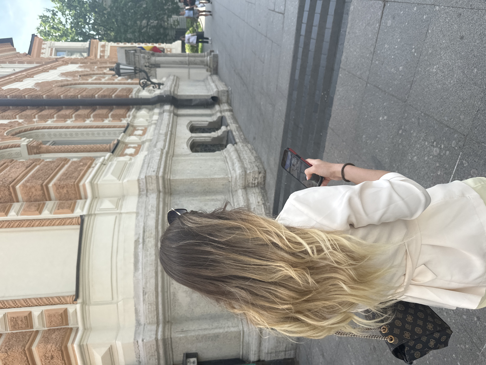 A woman with ombre blonde hair stands facing away from the camera, holding a smartphone in her right hand. She is wearing a white blazer with a belted waist and light yellow pants, and a black patterned handbag with a gold chain strap is slung over her left shoulder. The woman is standing on a paved city sidewalk in front of a large, ornate building with an orange and cream facade, featuring arched windows and decorative stonework. In the background, several other people are walking on the sidewalk, and a tree is visible to the right. The architecture suggests a location in the historic center of Saint Petersburg, Russia, likely near a landmark building. The lighting indicates it is daytime with a bright sky.