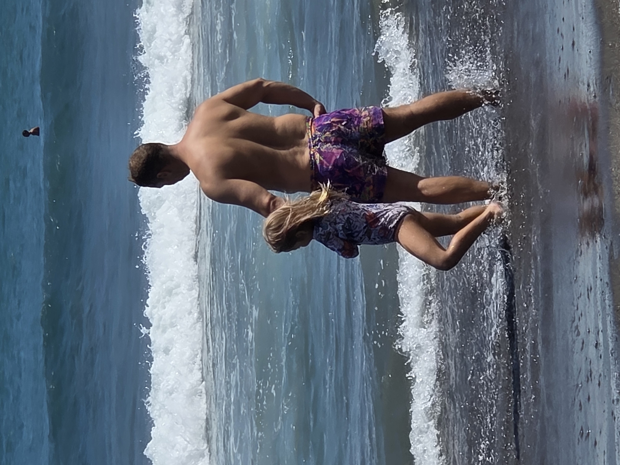 In North Kuta, Indonesia, a man and a young girl stand near the water's edge on a beach. The man, viewed from behind, wears colorful floral patterned swim trunks. The girl, also seen from behind and to his left, wears a patterned swimsuit. They are both looking out towards the ocean, where waves are breaking. In the distance, another person can be seen in the water. The water is a deep blue, and the sky appears clear and bright.
