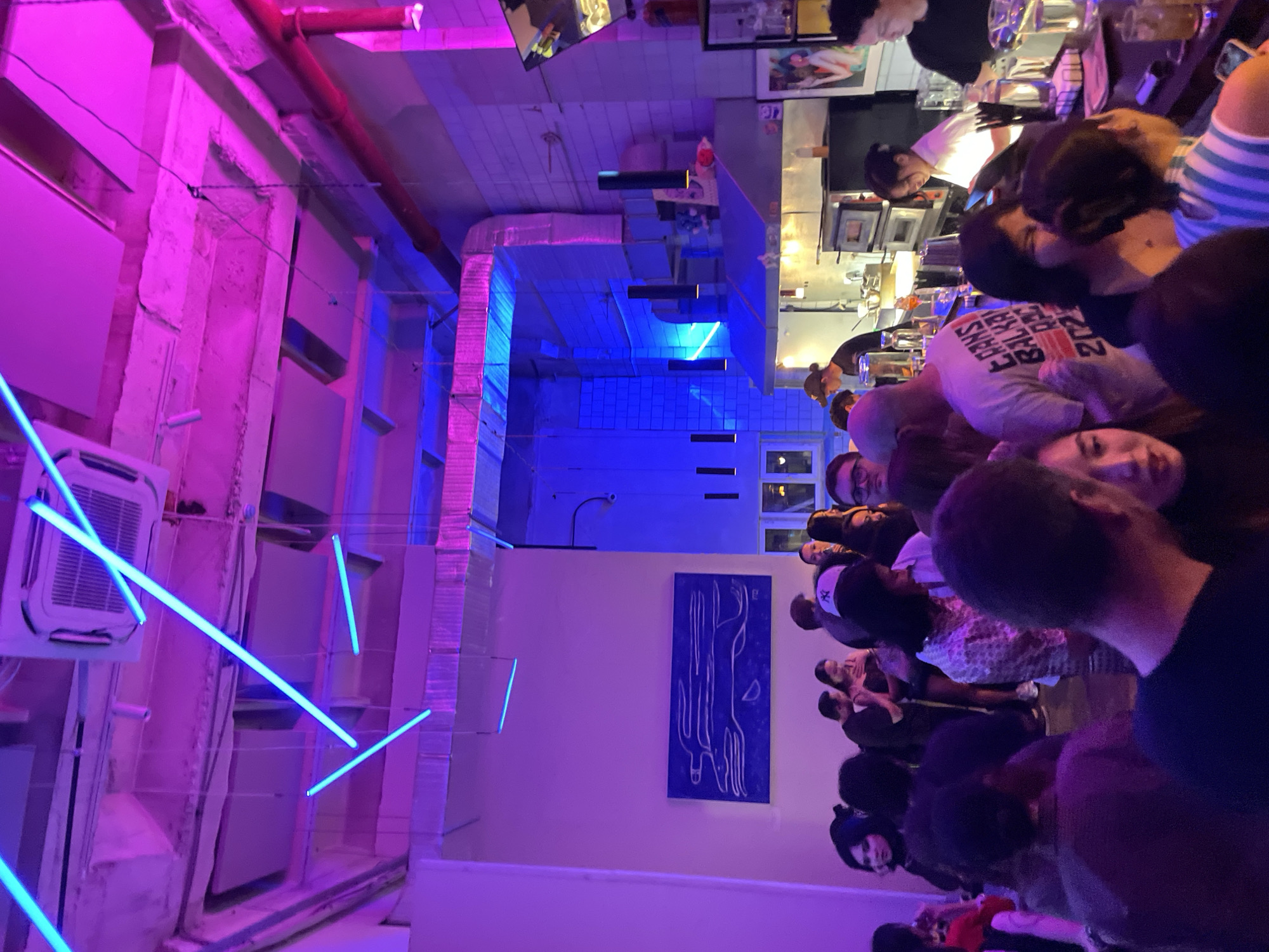 This is a medium-shot, indoor, low-angle photograph of a crowded bar or restaurant, possibly in Almaty, Kazakhstan, as indicated by the context. The scene is dimly lit and bathed in the neon glow of blue and pink lighting fixtures that crisscross the ceiling.
In the foreground, a diverse group of people are gathered, most of whom are facing away from the camera or are partially obscured, suggesting they are interacting with each other or the bar area. Some individuals appear to be in conversation, while others are simply standing or observing. The overall mood appears lively and social.
In the mid-ground, a bar or kitchen area is visible, featuring stainless steel appliances, including what appears to be a pizza oven, and shelves stocked with bottles and glasses. Several individuals are working behind the counter, preparing drinks or food. A prominent piece of blue artwork with white line drawings is displayed on a light-colored wall to the left of the frame.
The environment is characterized by industrial elements, such as exposed ductwork and concrete ceilings, contrasted with modern artistic lighting and décor. The atmosphere suggests a trendy, urban establishment.
