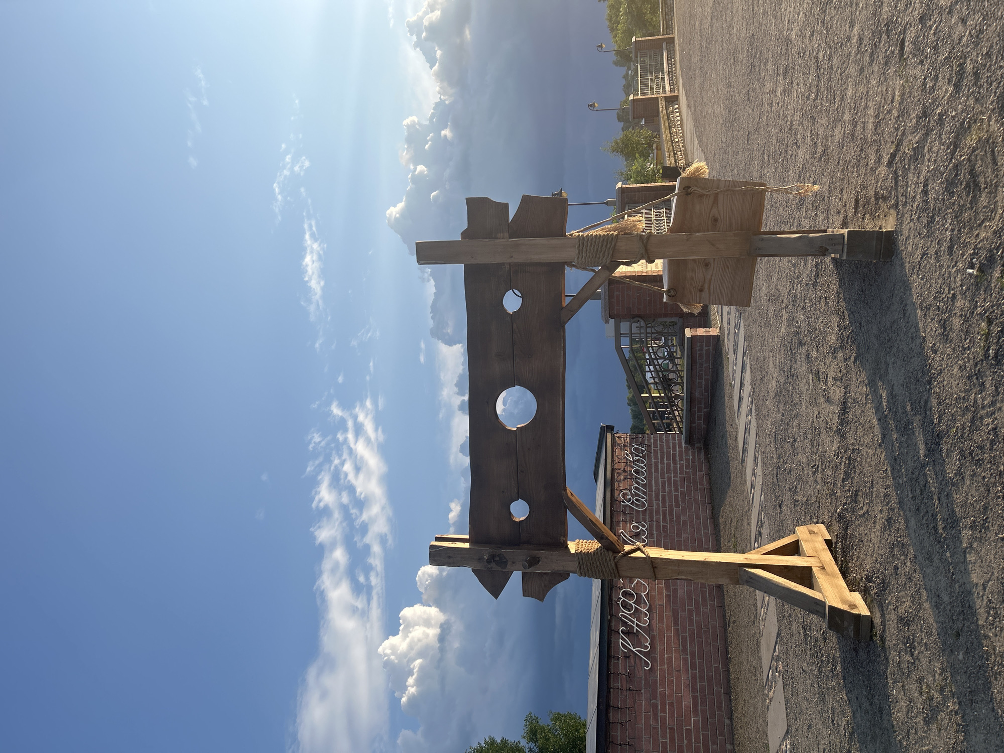 The image depicts an outdoor scene with a wooden stocks structure prominently featured in the foreground. The stocks are constructed from rough-hewn wood, with two circular openings for a person's head and hands, and a pointed arrow shape on one side. A small wooden sign with what appears to be fur trim hangs from a rope attached to the stocks. In the background, a brick wall with cursive lettering in white is visible, along with a metal fence and some streetlights. The ground is covered in gravel. The sky above is a vibrant blue with dramatic, fluffy white clouds and shafts of sunlight. The overall impression is a historical or themed display, possibly part of a park or recreational area in Zhukavichy rural council, Belarus. There are no people visible in the frame.