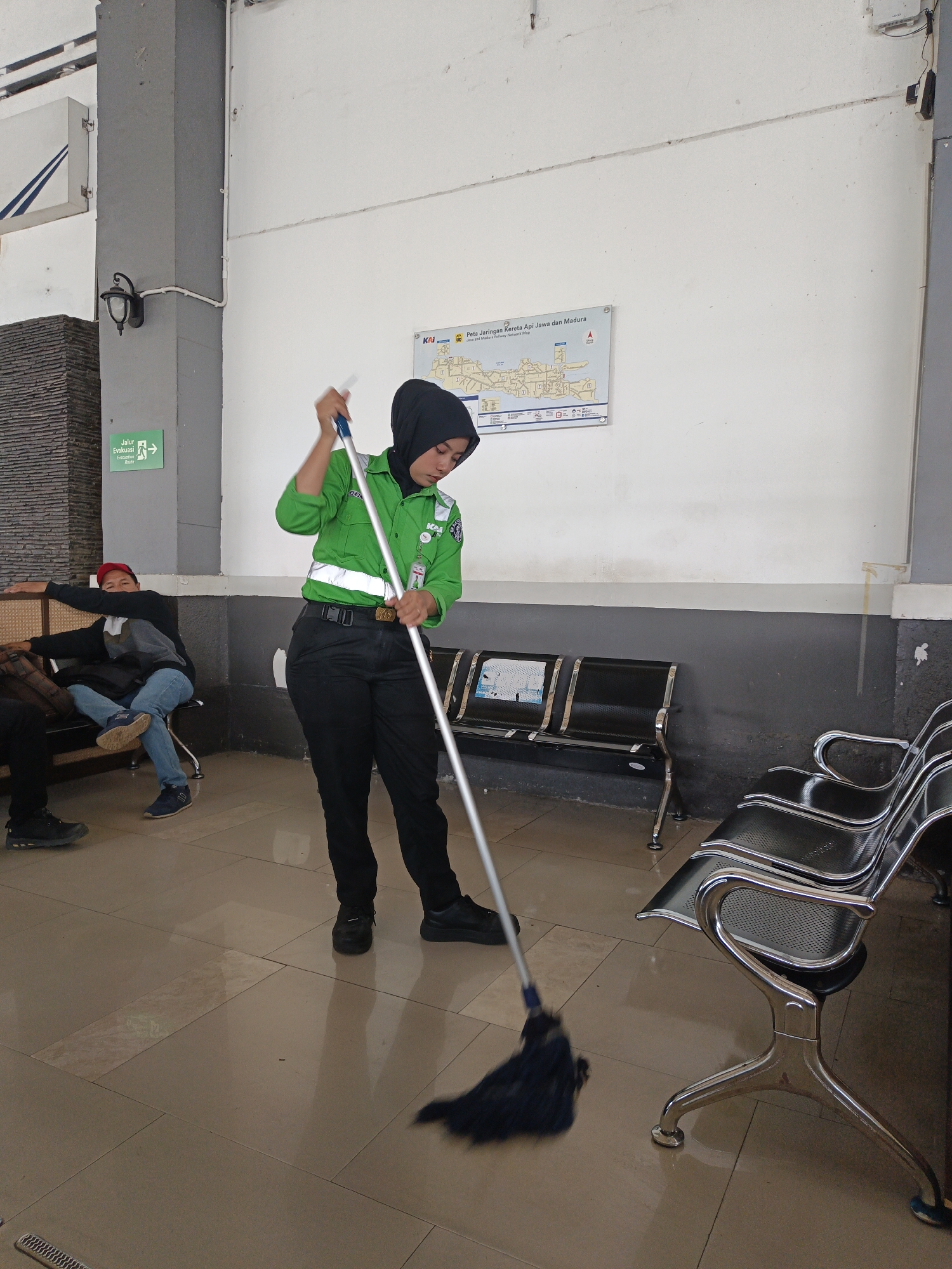 A woman wearing a green uniform and a black hijab is mopping a tiled floor in what appears to be a waiting area, likely at a train station in Klaten, Indonesia. She holds a mop with a blue head and a silver handle, actively cleaning. In the background, several rows of empty black metal chairs with silver frames are neatly arranged. A map labeled "Peta Jaringan Kereta Api Jawa dan Madura" (Railway Network Map of Java and Madura) is visible on the white wall. To the left, two men are seated, one wearing a red cap. A green "Jalur Evakuasi" (Evacuation Route) sign with an arrow is also present on the wall. The lighting suggests an indoor space with natural light filtering in.