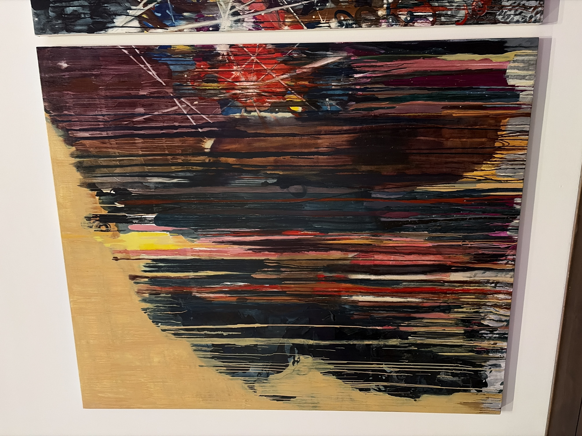 The image shows an abstract painting hanging on a white wall. The painting features horizontal bands of color, primarily in shades of brown, red, black, and yellow. The brushstrokes are textured and appear to be applied in a layered manner. The bottom left corner has a concentration of yellow and gold hues. There is another abstract painting partially visible above this one. The location is likely an art gallery or a private residence in Austin, United States of America.