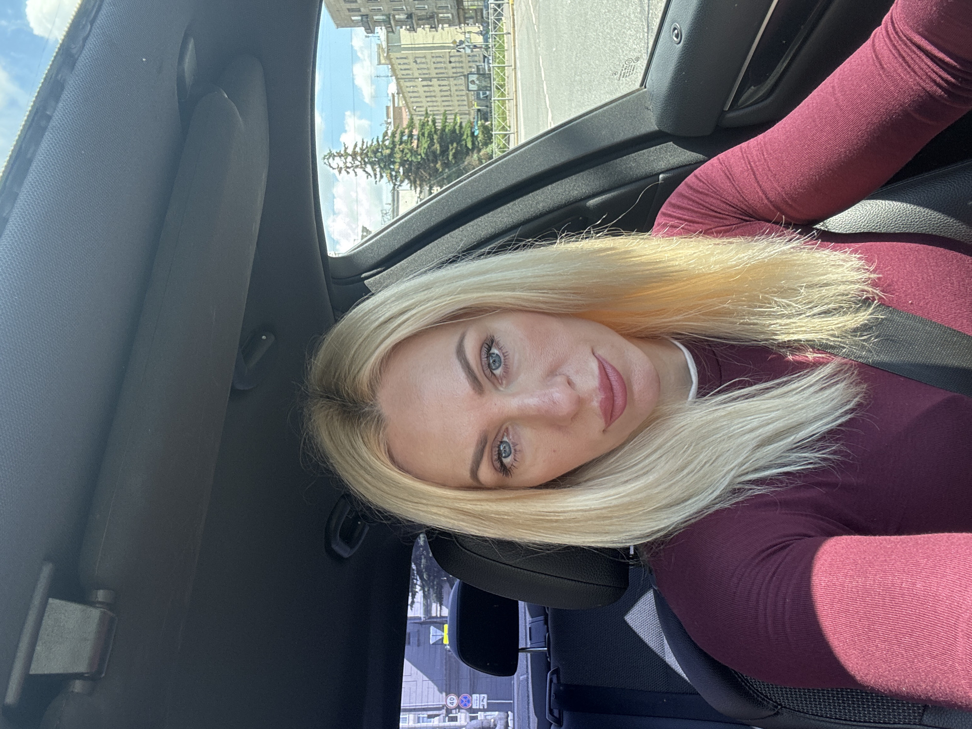 A blonde woman with blue eyes and pink lipstick is sitting in the driver's seat of a car. She is looking at the camera and smiling slightly. Her hair is long and straight and falls around her shoulders. She is wearing a maroon long-sleeved shirt. The interior of the car is visible, including the dashboard, steering wheel, and a seatbelt. The car appears to be in an urban environment, with buildings and a tree visible through the windows. The sky is blue with some clouds. The location is Saint Petersburg, Russia.