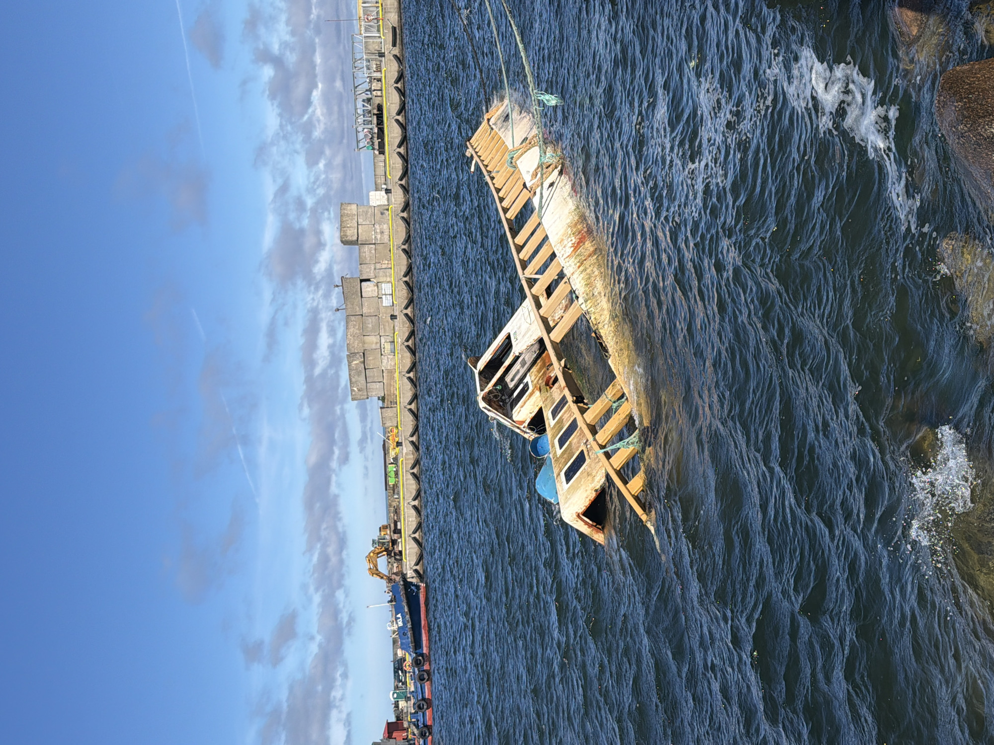 A partially submerged vessel, likely a small boat or barge, lists heavily in the dark blue, choppy waters of a harbor. The boat's wooden structure is visible, with some sections submerged and others above the waterline. Ropes are tied to the vessel, suggesting it may be secured or in the process of being salvaged. In the background, a larger blue and red vessel is docked, with an excavator visible on the shore behind it. The shoreline is lined with a concrete barrier and what appear to be storage containers or industrial structures. The sky is a bright blue with scattered clouds, indicating a clear day. The location is identified as the city of Viimsi, Estonia.