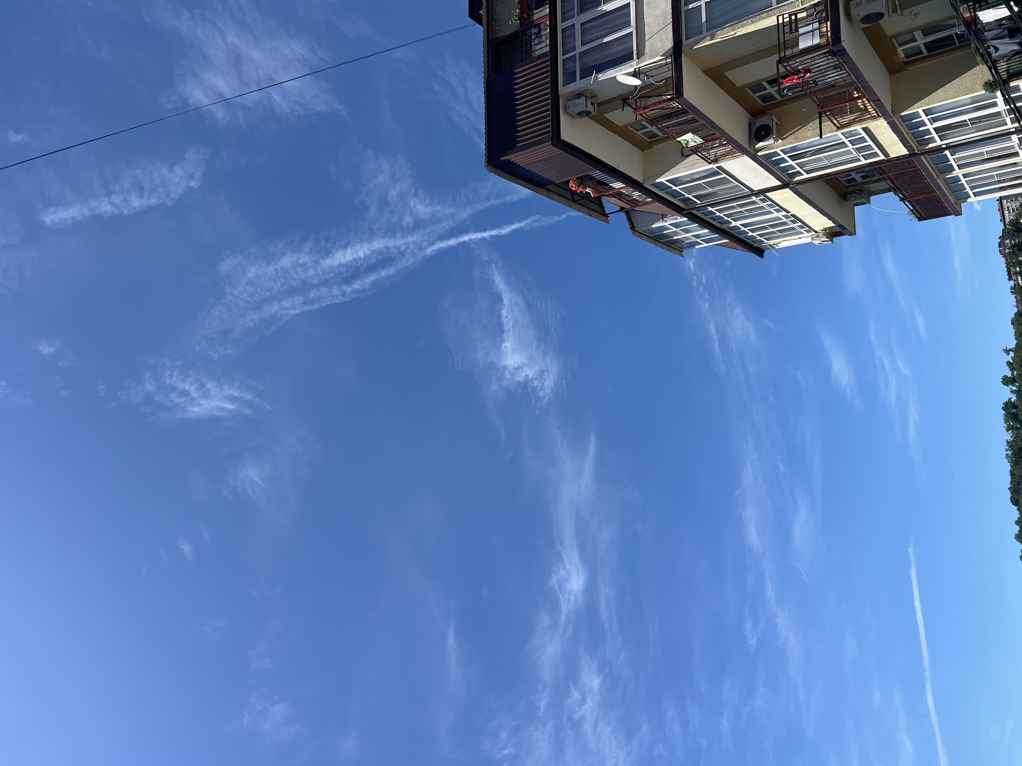 The image captures an upward view of a multi-story building under a bright blue sky. The building, located in Sochi, Russia, features a light yellow-beige facade with numerous rectangular windows and balconies. Many windows show internal blinds or curtains, and some balconies are adorned with small flowering plants in pots. The balconies have dark reddish-brown metal railings, and several external air conditioning units are visible on the building's walls. One balcony also features a white satellite dish. A dark corrugated metal roof section is visible on the upper left side of the building.
The sky above is clear and vibrant blue, scattered with thin, wispy white clouds, typical of cirrus formations. Several long, linear contrails from aircraft stretch across the sky, suggesting air traffic. A thin black wire or cable diagonally traverses the upper left portion of the frame. Along the right edge of the image, the tops of dense green trees are visible, indicating proximity to foliage or a park. The strong light suggests it is daytime, likely late morning or early afternoon, under fair weather conditions. No people or visible text are present in the image.