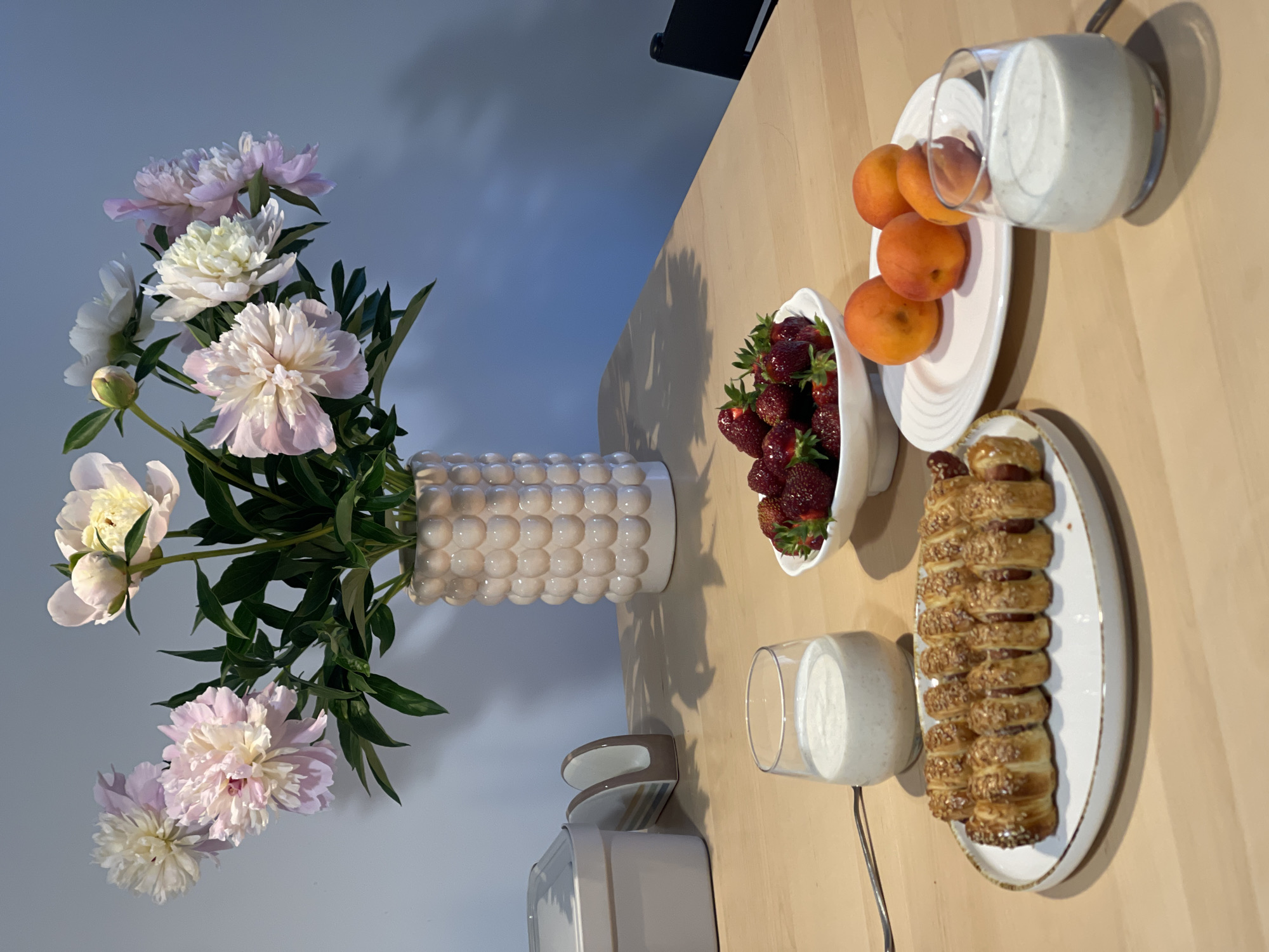 An overhead shot captures a wooden table set for a meal. A vase of pink and white peonies sits at the back, casting a shadow on the wall. In front of the vase, a cream-colored food container and a napkin holder are visible. A bowl of strawberries is centrally placed, to the right of which is a plate with four apricots. Two glasses of a creamy beverage are also on the table, one to the left and one to the right. A long pastry with sausages inside is presented on a plate to the left. The overall scene is clean and organized, with a focus on fresh food and floral decoration, set in an indoor environment that appears to be a dining area.