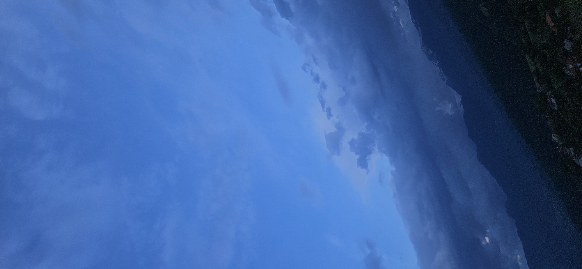The media file displays a panoramic vertical shot, primarily dominated by a vast sky over what appears to be a landscape in Gutsal, Bulgaria. The left side of the frame features a clear, light blue sky with faint, wispy cloud formations. As the view shifts towards the center and right, the sky transitions to a deeper, darker blue, heavily laden with more prominent, dense, and textured clouds, suggesting a significant cloud cover or an approaching weather system.
Along the far right edge of the image, a dark, undulating landmass, likely a mountain range or hills, is visible. This terrain appears silhouetted against the darker sky, with indistinct features due to low light. Nestled on this landmass, particularly in the lower right corner, are faint outlines of what appear to be scattered buildings or a small village.
The overall impression is that of twilight, either early morning before sunrise or late evening after sunset, given the deep blue hues and the absence of direct sunlight. The presence of both clear sky and heavy clouds indicates dynamic weather conditions. No specific activity or event is occurring, and there are no visible people, distinct objects beyond the natural landscape, or text in the image.