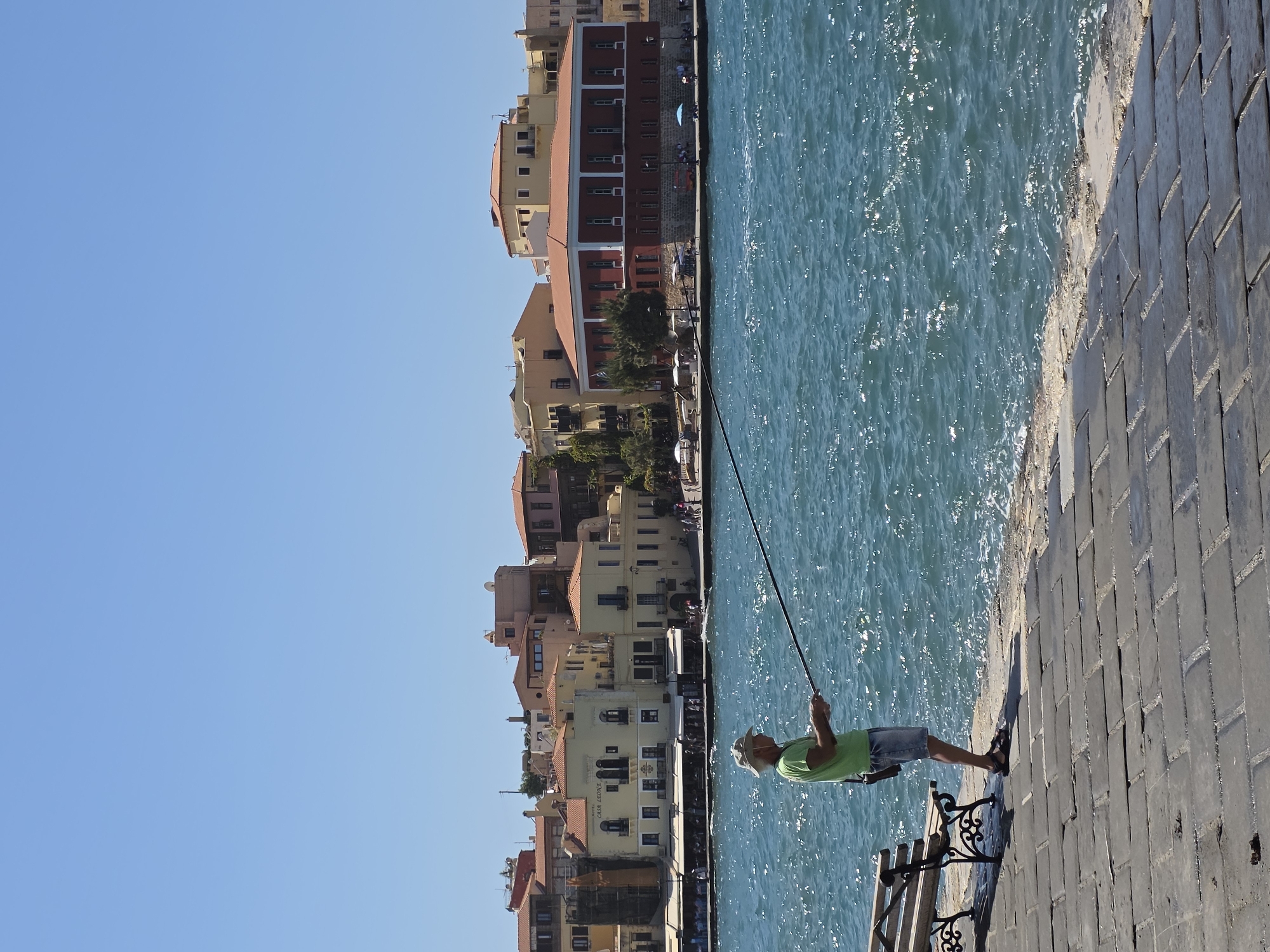 A man fishes on a sunny day in the old harbor of Chania, Greece. He stands on a paved walkway made of large, rectangular stones, facing the sparkling turquoise water. The man wears a light-colored hat, a green t-shirt, and denim shorts. He holds a fishing rod, its line extending into the sea. To his left, a wooden bench with ornate black metal legs sits on the walkway. In the background, a waterfront promenade is lined with buildings featuring red-tiled roofs and light-colored facades, indicative of Venetian and Ottoman architecture. The sky above is a clear, bright blue.