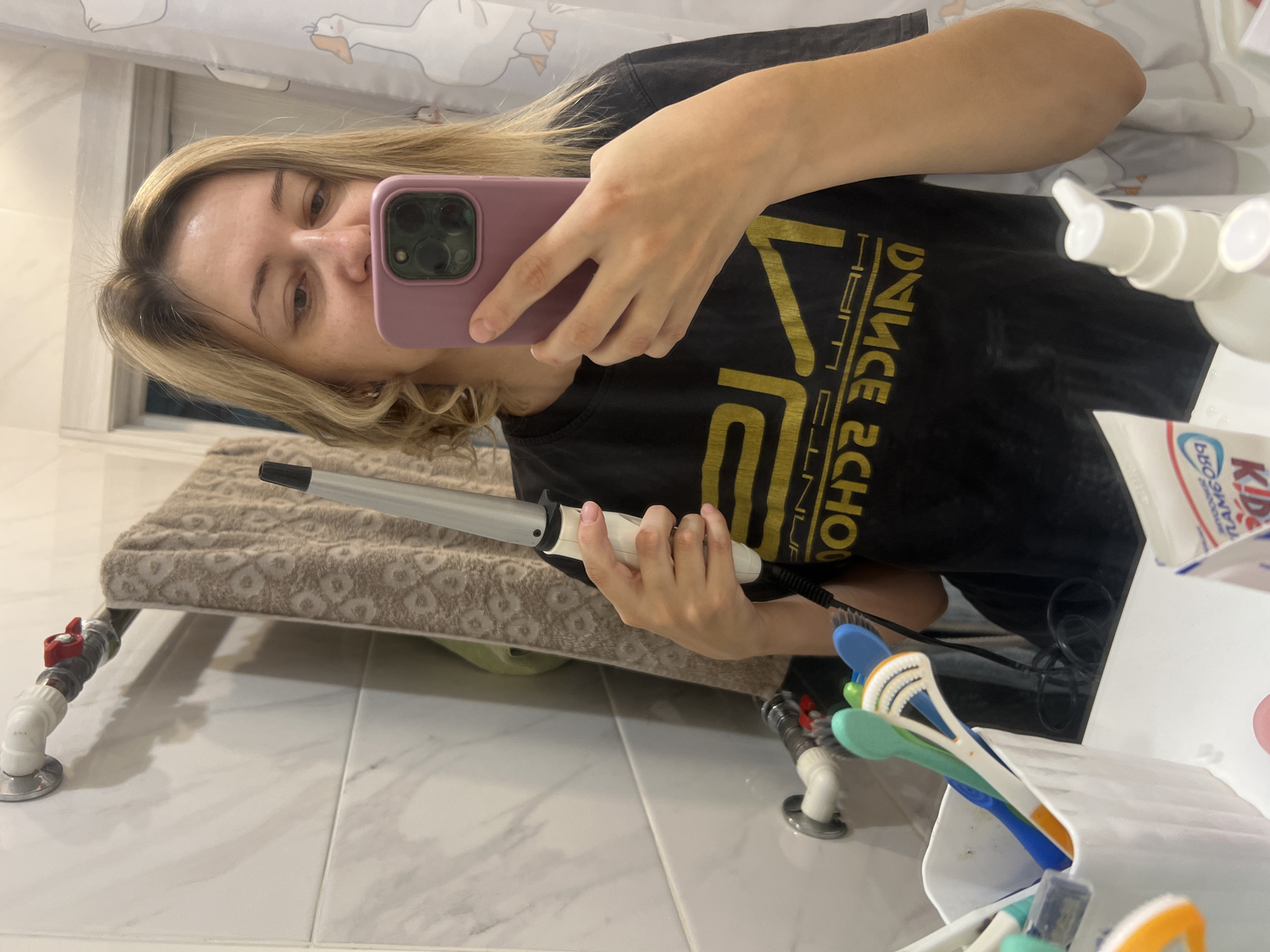 A woman with blonde hair is taking a selfie in a bathroom mirror. She is holding a curling iron in her right hand and a pink phone in her left hand. She is wearing a black t-shirt with yellow text. In the background, there is a towel hanging on a rack, a shower curtain with a duck pattern, and a sink with toothbrushes and toothpaste. The scene appears to be indoors, possibly in a home or hotel bathroom. The image does not contain any information that would allow for a determination of its location in Volgograd, Russia.