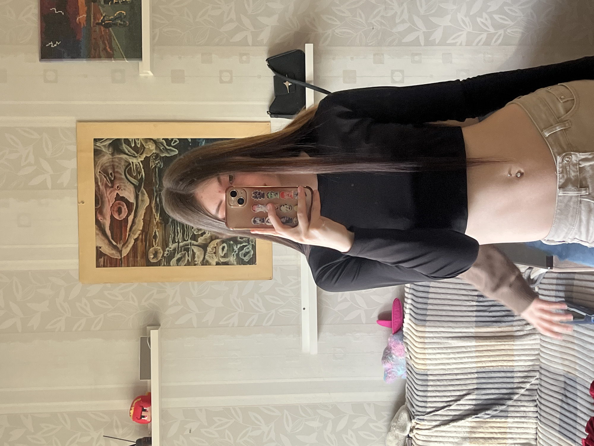 A young woman is standing indoors, holding a smartphone in front of her face to take a selfie. She is wearing a black long-sleeved crop top and light-colored jeans. Her midriff is exposed, and a navel piercing is visible. The background includes a beige wallpaper with a leaf pattern, a framed painting of a grotesque face, another framed painting, and a shelf with a small black purse. A striped blanket is draped over a bed or sofa. The location is Unknown, Russia.