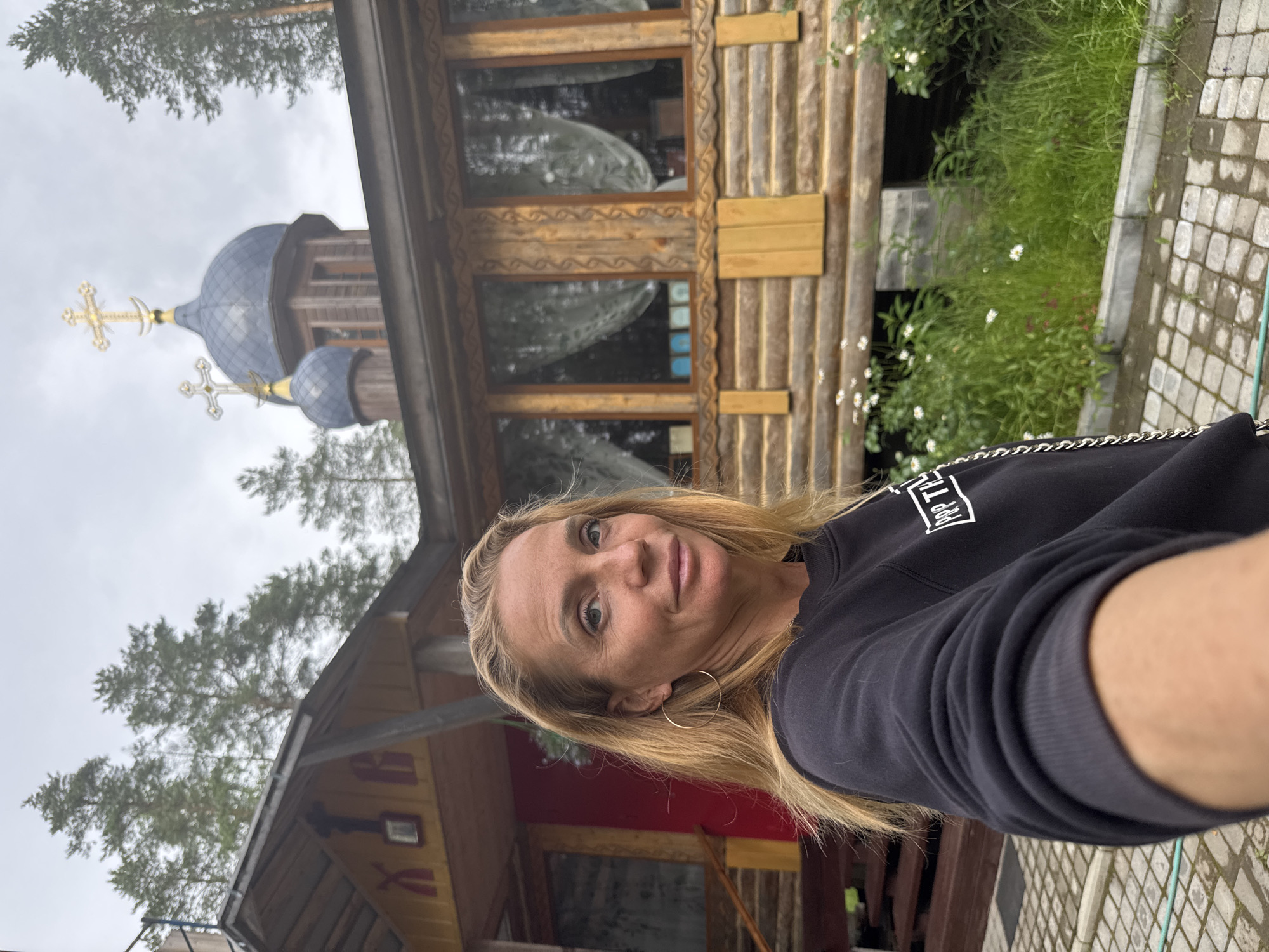 A blonde woman with hoop earrings, wearing a black sweatshirt, is taking a selfie outdoors. Behind her is a wooden building with two blue domes topped with crosses, suggesting it is a church or monastery. The building has a red section and wooden log construction. The environment appears to be rural, with trees and green vegetation surrounding the structure. White flowers are visible in the foreground. The sky is overcast. The location is identified as Neniämäki, Russia.