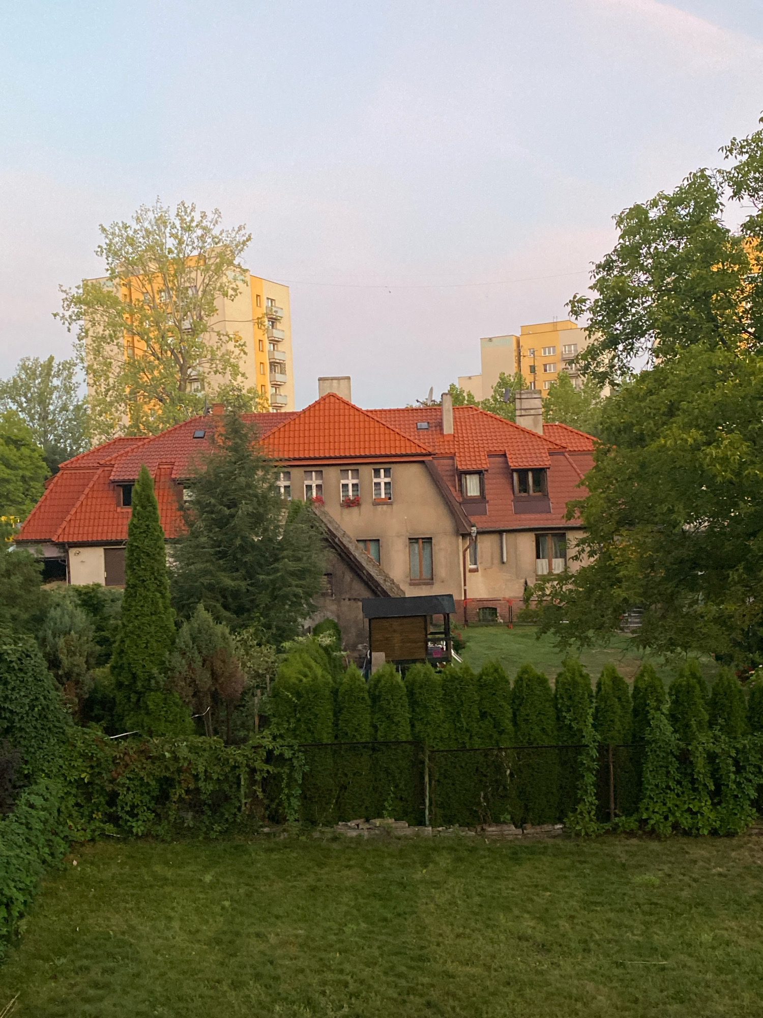 The image shows a two-story house with a red tiled roof, surrounded by trees and vegetation. The house has a light-colored facade with several windows. In the background, two tall apartment buildings are visible, suggesting an urban or suburban setting. The foreground features a well-maintained lawn and a fence enclosing a garden area. The sky is a pale blue, indicating it is likely daytime, possibly early morning or late afternoon due to the soft lighting. The overall scene depicts a residential property with a blend of natural and built environments. The presence of distinct architectural styles of the house and apartment buildings, along with the visible vegetation, points to a European city, and more specifically, to a neighborhood within Katowice, Poland, characterized by its mix of older residential structures and more modern high-rise apartments.