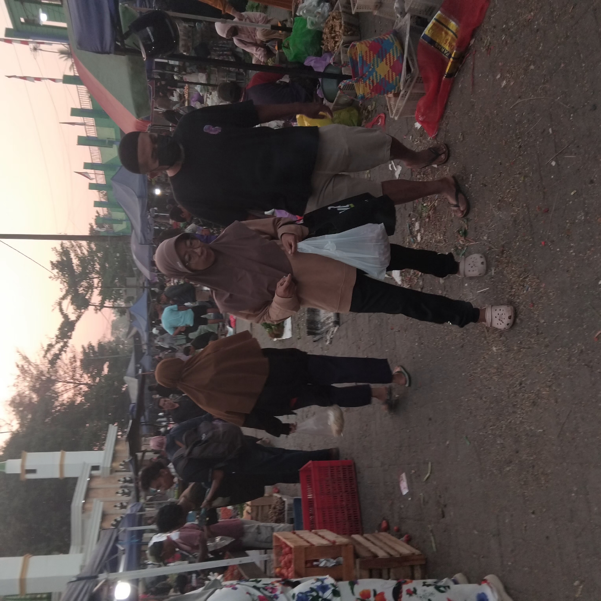 This is a medium-shot, eye-level, full-color photograph taken outdoors in what appears to be a market setting. The time of day seems to be late afternoon or early evening, as indicated by the warm, diffused light and the soft, rosy hue of the sky. The ground is paved and appears somewhat dusty and littered with small debris.
In the foreground, a person wearing black pants and light-colored clogs is walking towards the right of the frame. They are carrying a translucent white plastic bag filled with items and a black tote bag. To their left, another person is partially visible, wearing a brown hijab and black pants, with their back to the camera. Further to the left, a stack of wooden crates is visible, one containing red produce, likely tomatoes. A red plastic crate is also present.
In the mid-ground, several people are interacting and browsing at stalls under awnings. Some are wearing headscarves, suggesting a cultural context common in parts of Indonesia. One man in the mid-ground, facing left, wears a black t-shirt, light-colored shorts, and a black face mask, along with a black skullcap. He is looking towards the left. Behind him, other individuals are visible, some with backpacks.
In the background, a prominent white and gold building with a green-topped minaret is visible, likely a mosque. Trees with dark foliage line the area. Several colorful awnings and market stalls are set up, some with lights visible, indicating ongoing commerce. In the far background, a green and white structure with red and white flags is present. The overall scene depicts a bustling outdoor market, likely in the city of Banjarbaru, Indonesia, with a mix of shoppers and vendors under the ambient light of dusk.