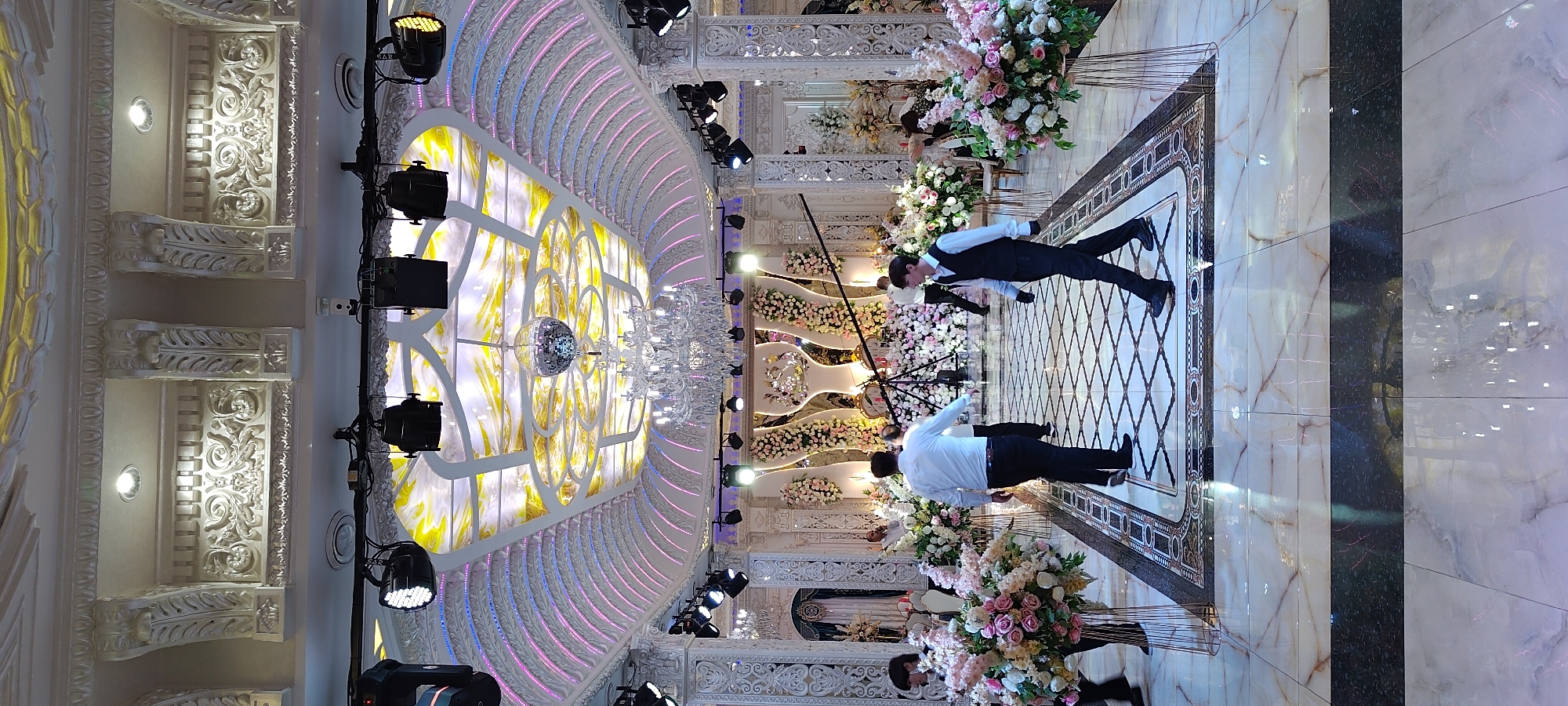 The image captures the interior of a lavish event hall in Khujand, Tajikistan. The hall features a highly ornate ceiling with intricate white stucco moldings, accented by yellow and white patterned panels. A large, crystal chandelier hangs prominently, and a disco ball is suspended above it, indicating a celebratory atmosphere. The walls are decorated with elaborate white relief carvings and illuminated by pink and blue accent lights lining the curved ceiling structures.
The floor is a polished marble with intricate geometric patterns in a diamond motif, bordered by darker marble with decorative inlays. Large floral arrangements with pink and white flowers are strategically placed throughout the hall. Several people are present, some of whom appear to be staff or event personnel. In the foreground, two individuals in formal attire, likely a waiter and a guest or another staff member, are walking across the floor. The waiter is dressed in a white shirt and black vest, while the other person wears a white shirt and black trousers. Other individuals are visible in the background, attending to preparations or engaged in conversation. The overall scene suggests a high-end event, possibly a wedding or a formal reception, set in a well-decorated and luxurious venue.