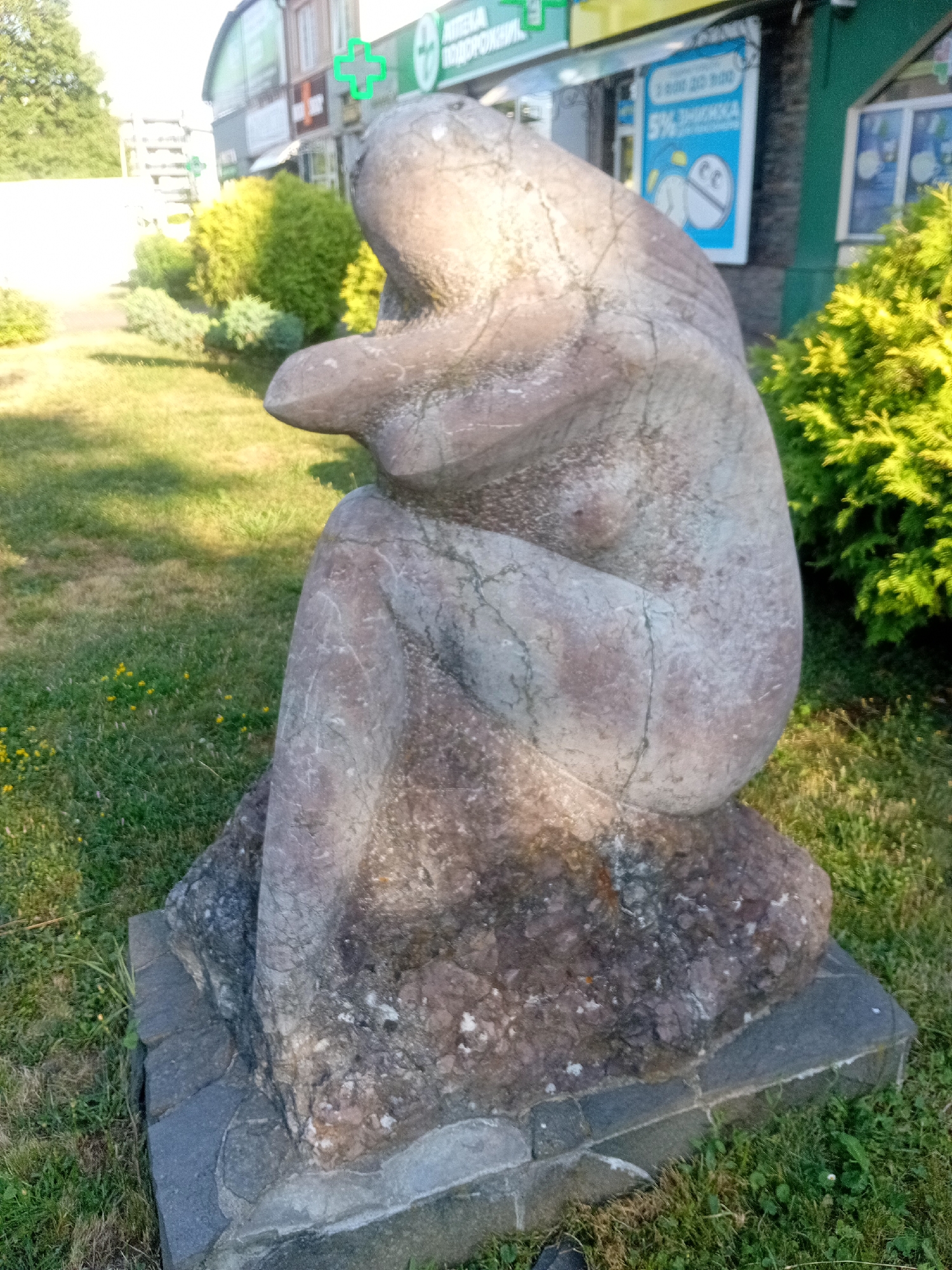 An outdoor, eye-level, medium shot captures a stone sculpture of a seated, nude female figure in the foreground. The sculpture is positioned on a grey stone pedestal and is set against a backdrop of green grass and foliage. Behind the sculpture, to the left, a paved walkway is visible, leading towards a building. To the right of the sculpture, dense green bushes are prominent. Further in the background, a building with signage is visible, with green cross symbols indicating a pharmacy. The lighting suggests it is daytime, with sunlight illuminating the scene.