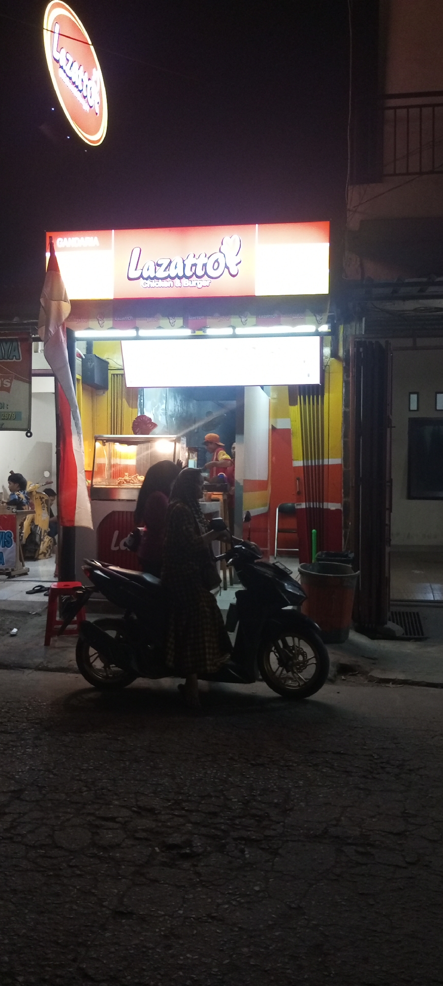 A nighttime outdoor scene in Setu, Indonesia, features a "Lazatto! Chicken & Burger" storefront illuminated by bright signage. The main sign above the shop reads "Lazatto!" in stylized white and pink lettering on an orange background, with smaller text below indicating "Chicken & Burger." A larger, oval-shaped neon sign with similar "Lazatto!" lettering is also visible hanging higher up.
In front of the shop, two women are standing next to a dark-colored motorcycle. One woman, wearing a patterned dress and headscarf, is seated on the motorcycle, while the other woman stands behind her. Behind the women and motorcycle is a food stall with a glass display case showcasing fried chicken and a food preparation area where a person in an orange cap and red vest appears to be working. A red stool is placed near the food stall.
To the left of the scene, a portion of another sign is visible with Indonesian text that reads "GANDARIA." A red and white Indonesian flag is prominently displayed on a pole to the left of the entrance. The ground in front of the shop is paved and appears somewhat cracked and uneven. A large orange bucket and a green trash can are situated to the right of the food stall. The overall lighting suggests it is evening or night, with the bright signs casting light on the immediate area.