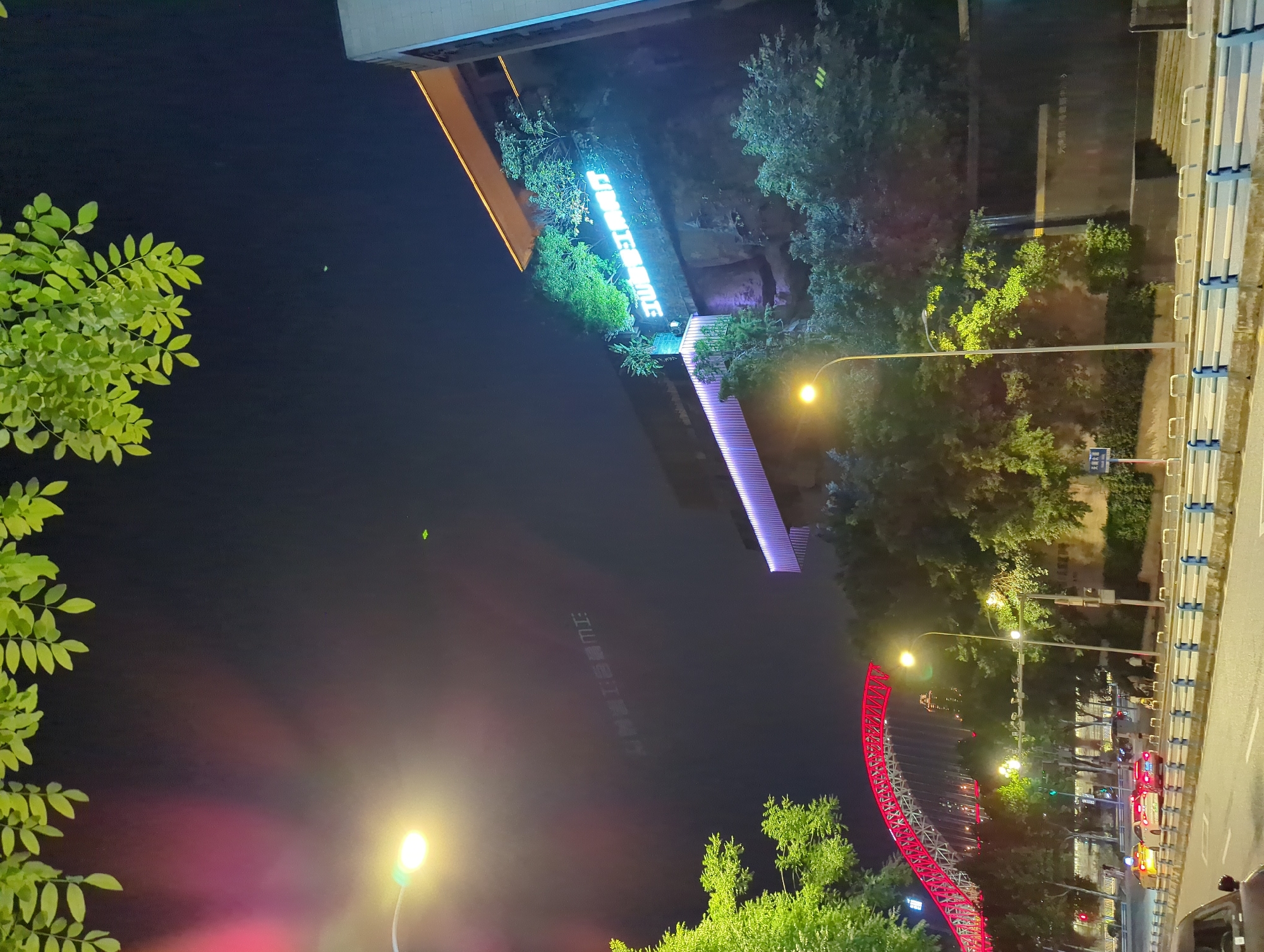 The image captures a nighttime urban scene in Liangjiang, China. A red illuminated bridge structure is visible in the mid-ground, arching over a street. On the right side of the image, a building is built into a hillside, with illuminated signage that appears to read "江景湾" (Jiāng Jǐng Wān). Green foliage overhangs the scene from the top. Streetlights illuminate the road, on which several cars, including taxis, are present. Pedestrians are seen on the sidewalk in the mid-ground. The overall impression is of a well-lit, developed urban environment at night.