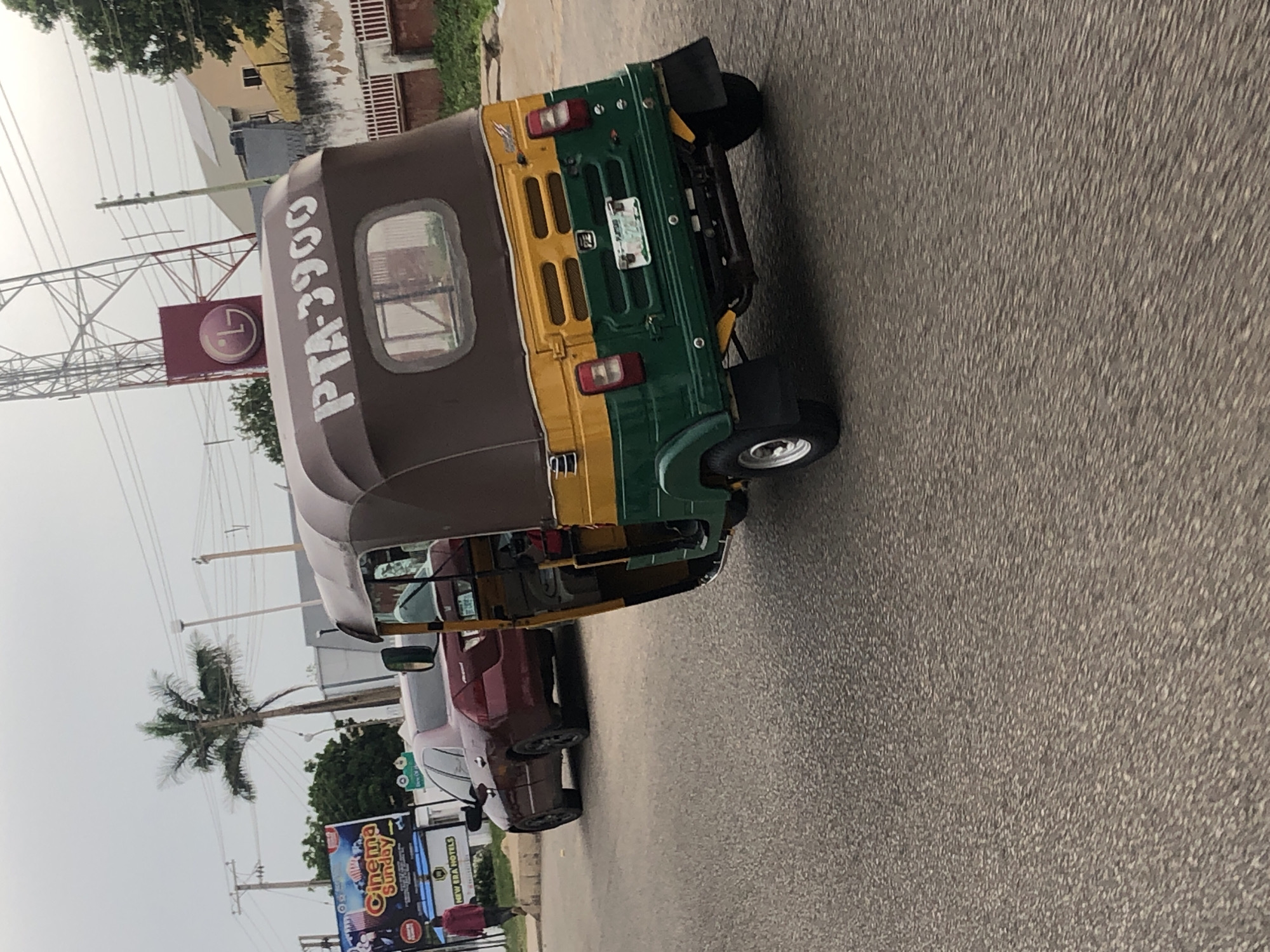 A three-wheeled auto-rickshaw with a brown and yellow canvas roof and green lower body is angled in the foreground, its license plate reading PTA-3900. A maroon sedan is visible in the background, and a person in a red jacket walks away from the camera. A palm tree and a billboard are also present, and overhead power lines and a telecommunications tower are visible. The scene appears to be a street in an urban environment.