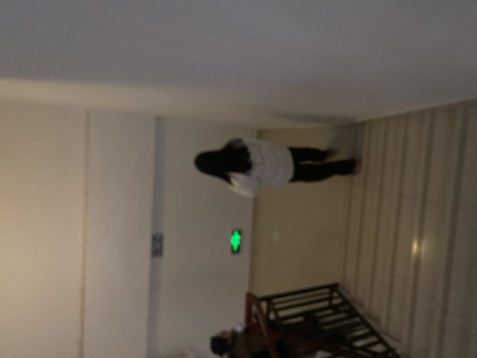A blurred and rotated view of a room features a person, seen from behind, wearing a white shirt and dark pants. The person is positioned horizontally in the frame. A door with a green illuminated figure is partially visible on the left. Beneath the person, a dark-colored bunk bed frame is situated. Striped wallpaper adorns the right side of the room. The background is out of focus.